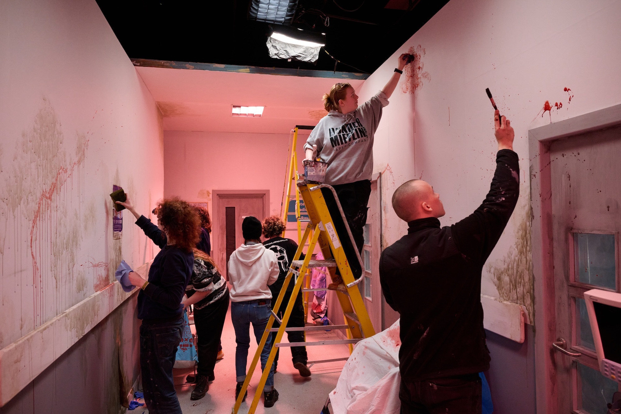 Group of Arts University Bournemouth students painting pink walls in a room with black ceiling, using brushes and rollers, yellow ladder visible.