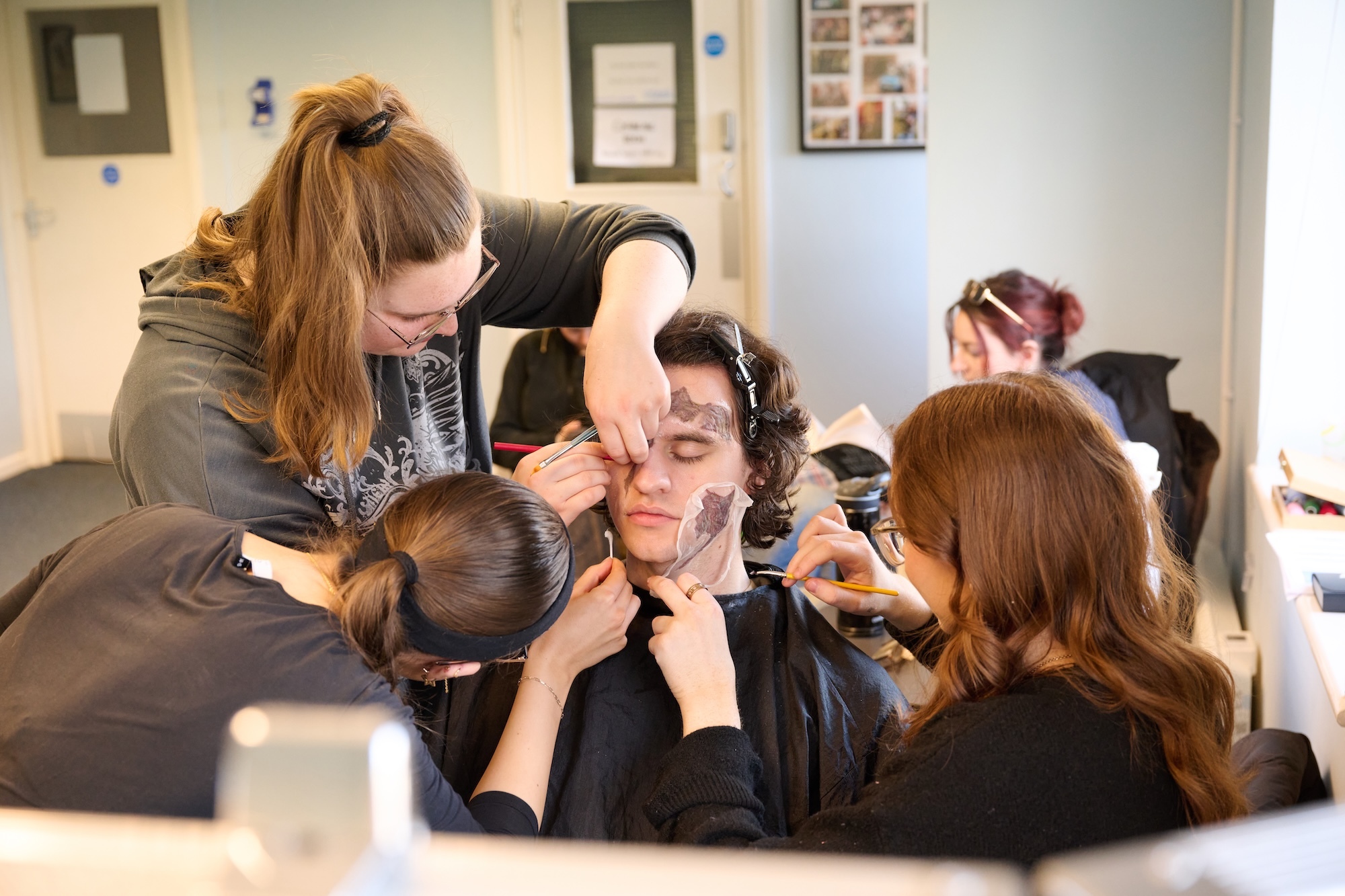 Actor J Walters in black smock having theatrical make-up applied by three BA (Hons) Make-up for Media and Performance students in bright studio with white walls and framed pictures.
