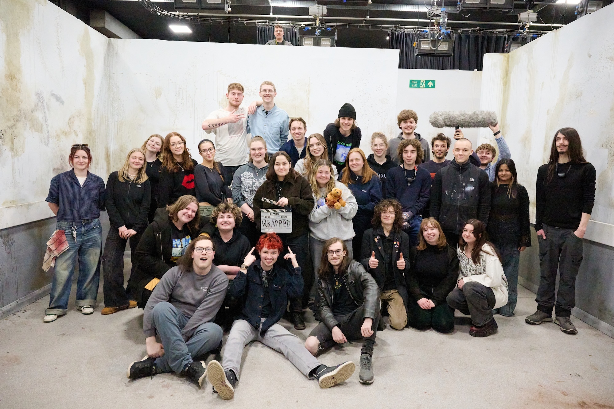 Large group of BA (Hons) Film Production students posing on a film set with industrial ceiling and green exit sign visible above.