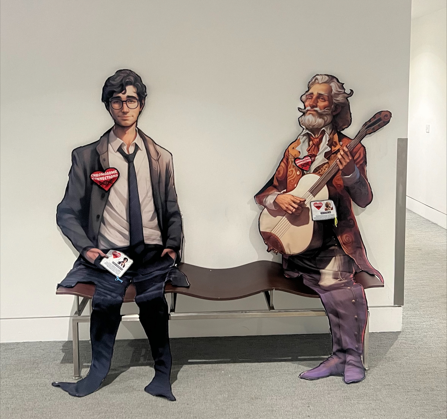Two cardboard cut-outs of men, both wearing heart-shaped badges, one wearing a suit and tie with glasses, the other in Victorian-style dress with a full beard playing a guitar. Both have fabric trousers and shoes folded over a bench as if sitting.