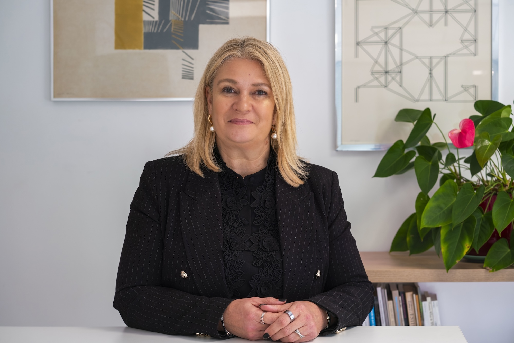 Blonde woman in black blazer sits at desk with framed artwork on wall behind her and green plant with red flower to right.