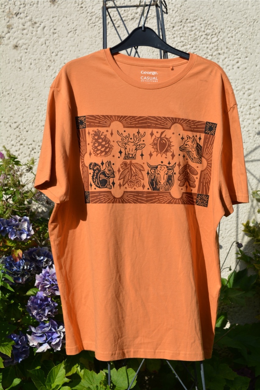 Orange t-shirt with printed black and red screen print designs, forest animals and leaves with a sunny border, hung up in a sunny garden.