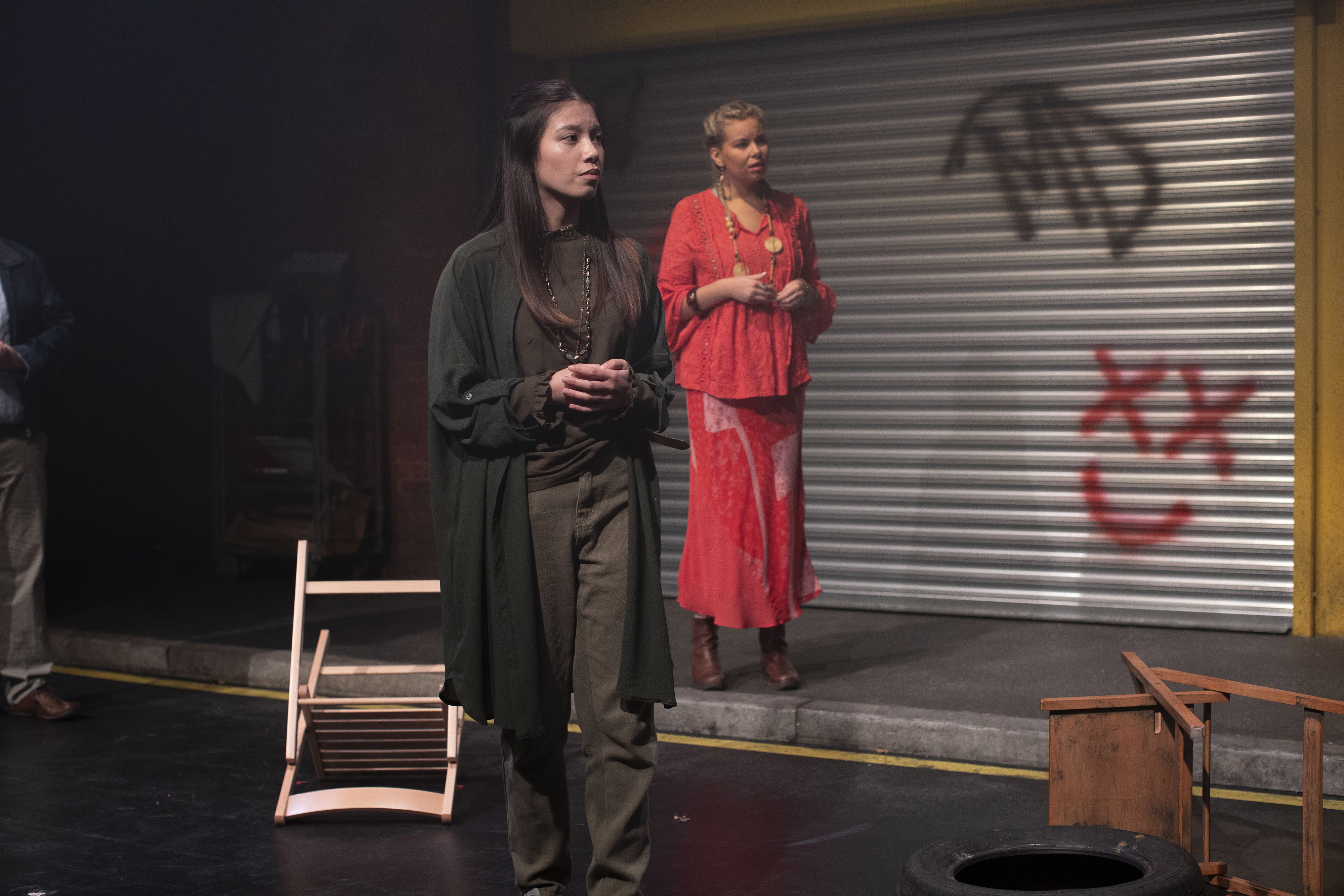 Two actors in costume on stage in front of a metal gate with graffiti on it.