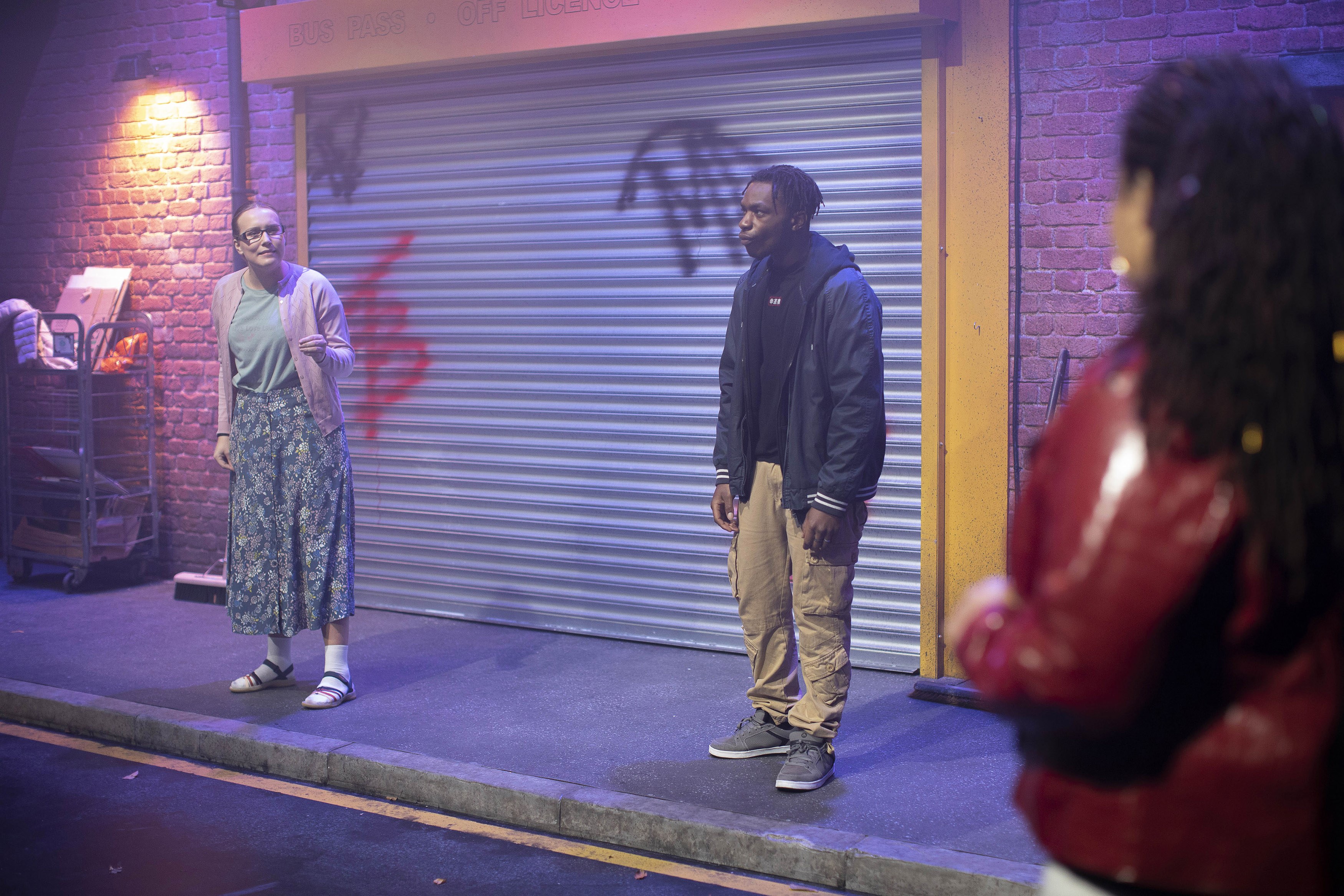 Three performers on a stage set on a street with a pavement. There is an off-license shop in the background with its metal shutters down.