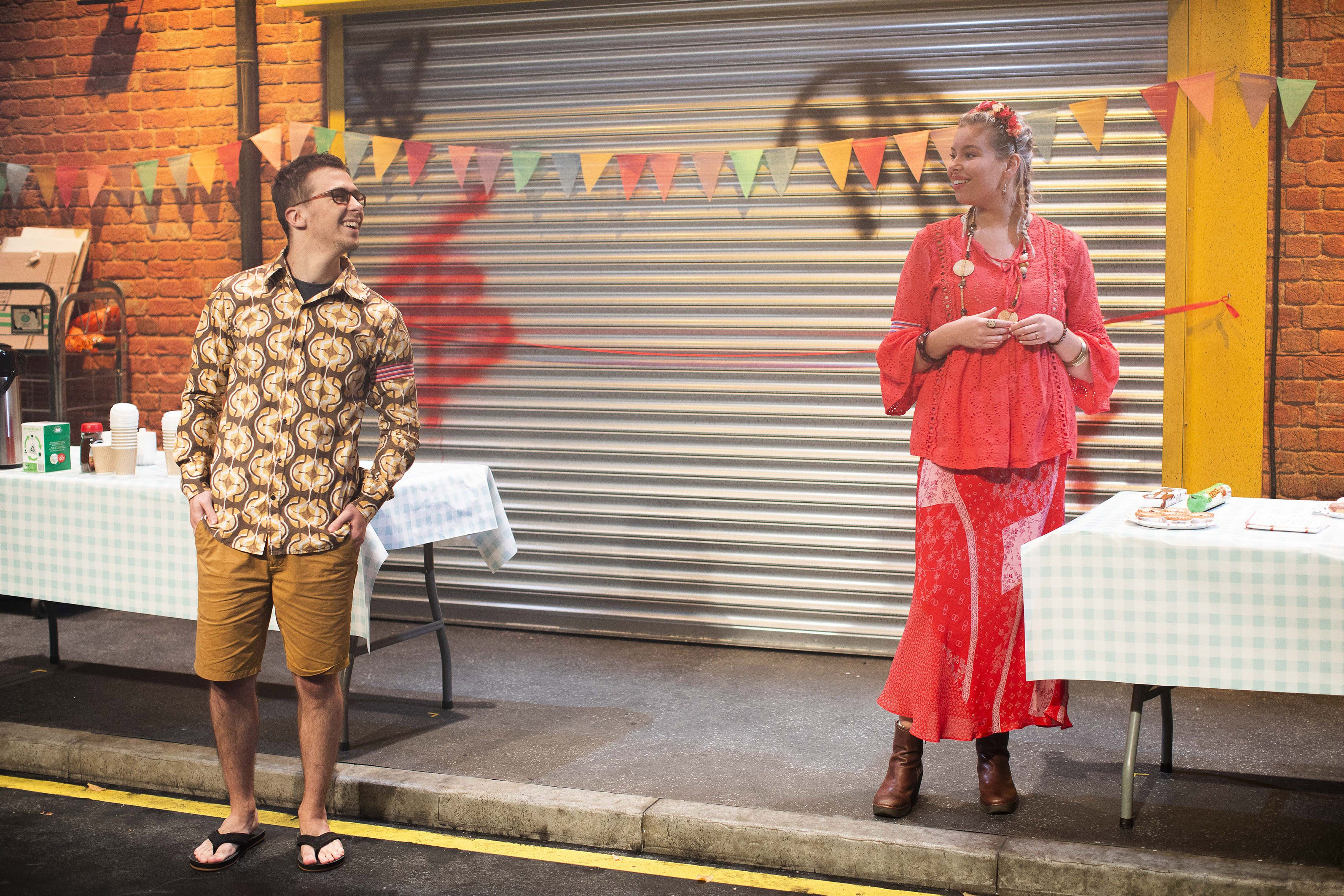 Two people on a stage set to look like a street party, with tables of food either side and bunting hanging in the background.