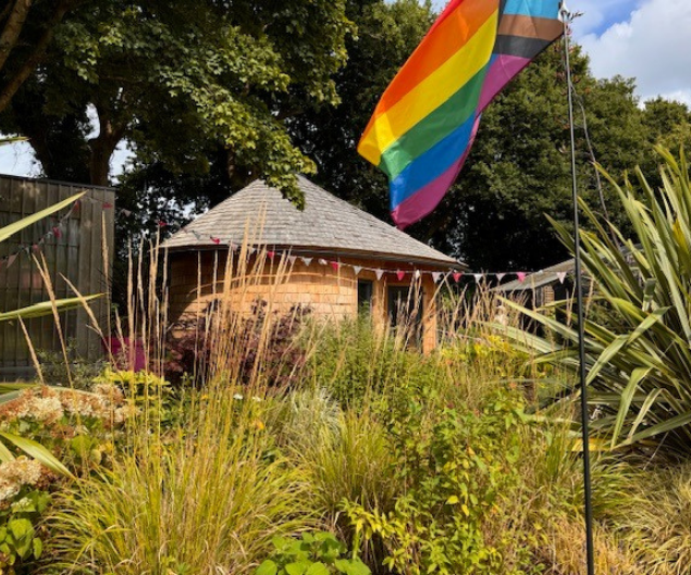 Small hut with round roof behind a garden area, LGBTQ+ flag and trees.