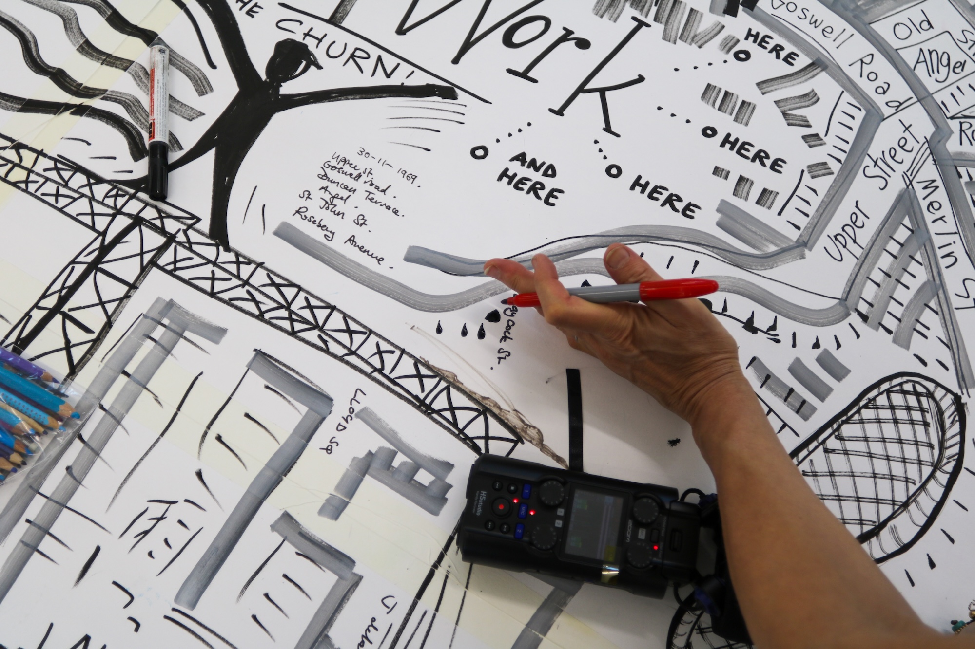 A hand holding a red pen over a black-and-white illustrated map or site plan, with a portable audio recorder and architectural sketches on the table.