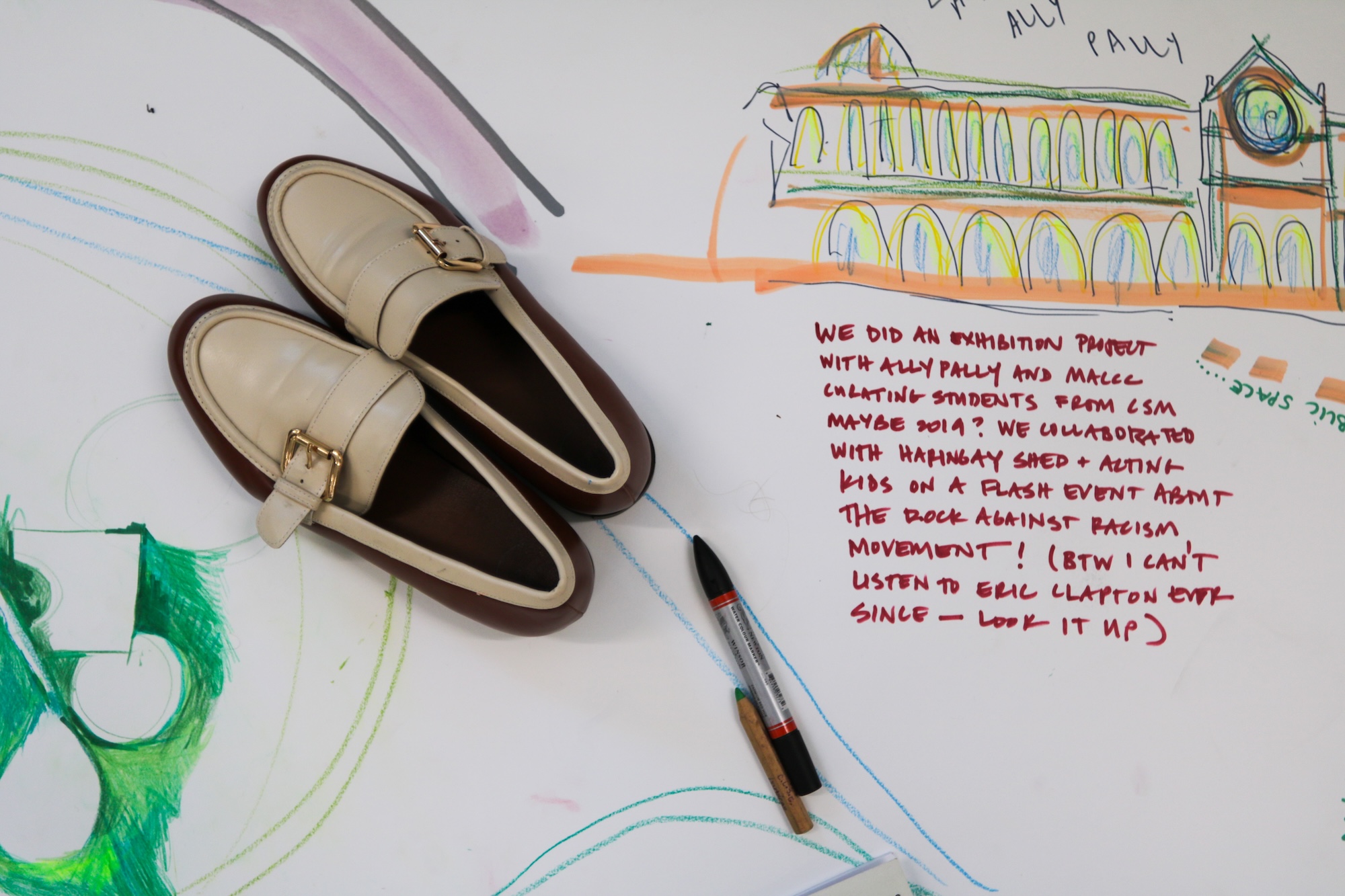 A pair of beige loafers with dark interiors lies on a white surface covered with hand-drawn sketches and notes, with a pen nearby.