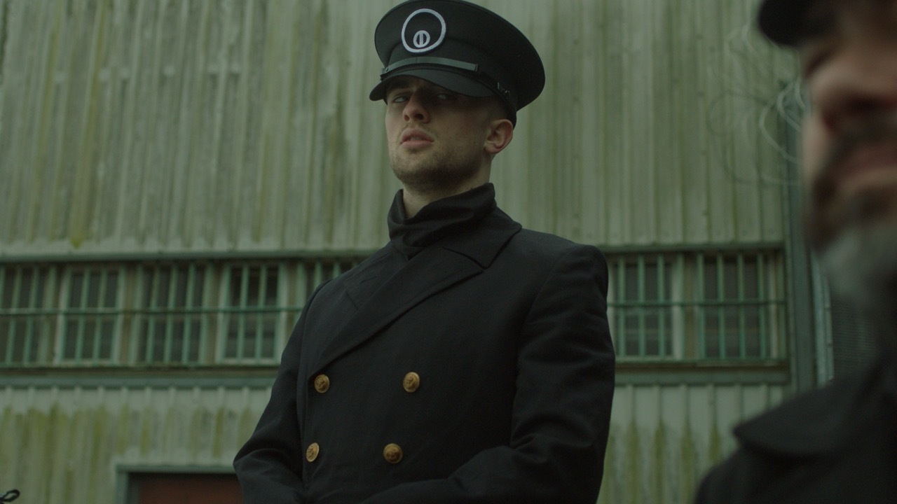 Man in dark naval uniform with peaked cap and gold buttons stands before green corrugated metal building.