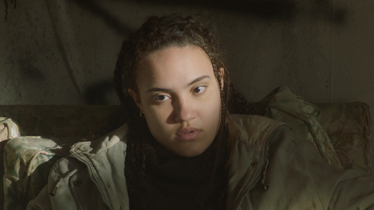 Woman with dark curly hair lying on bed, looking directly at camera in dimly lit room with muted brown and green tones.