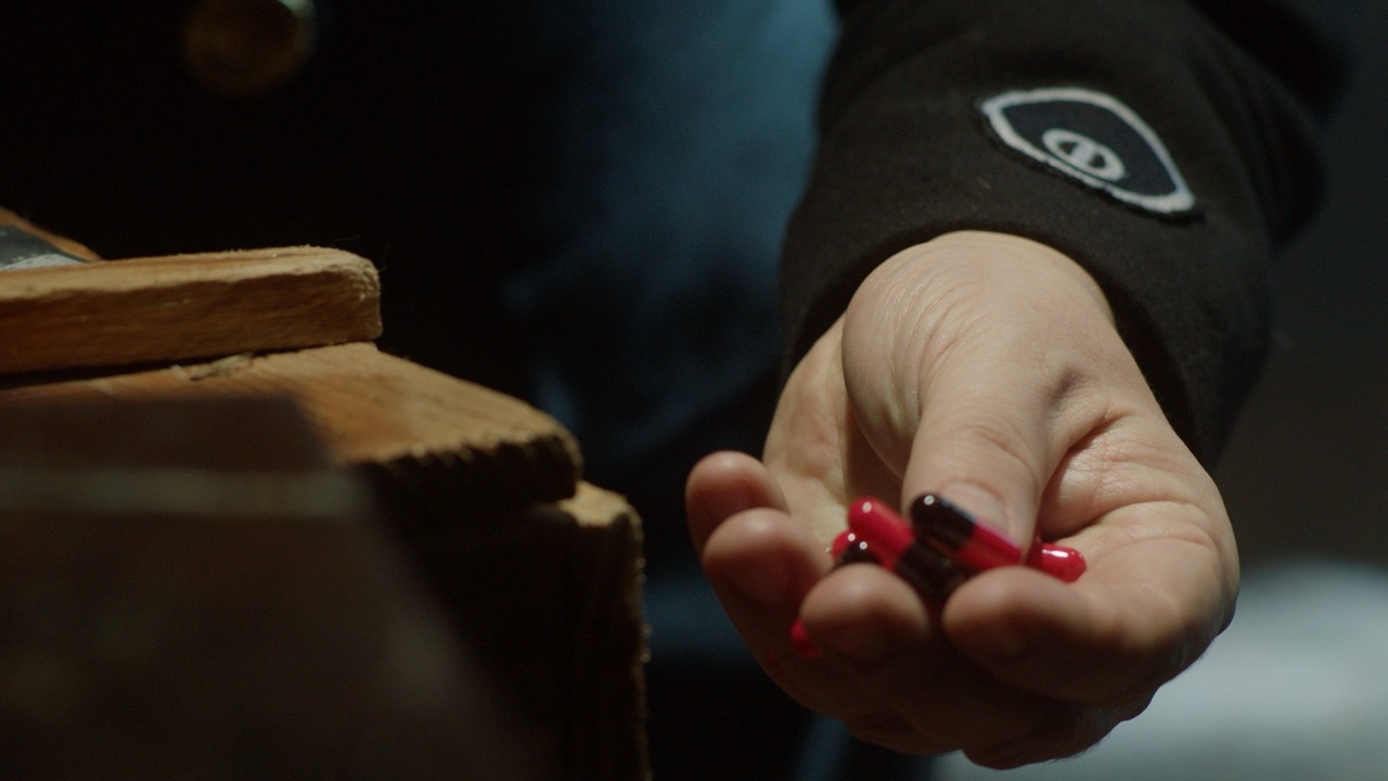 Open palm holding two red pills, with wooden boxes visible to the left, blurred background.