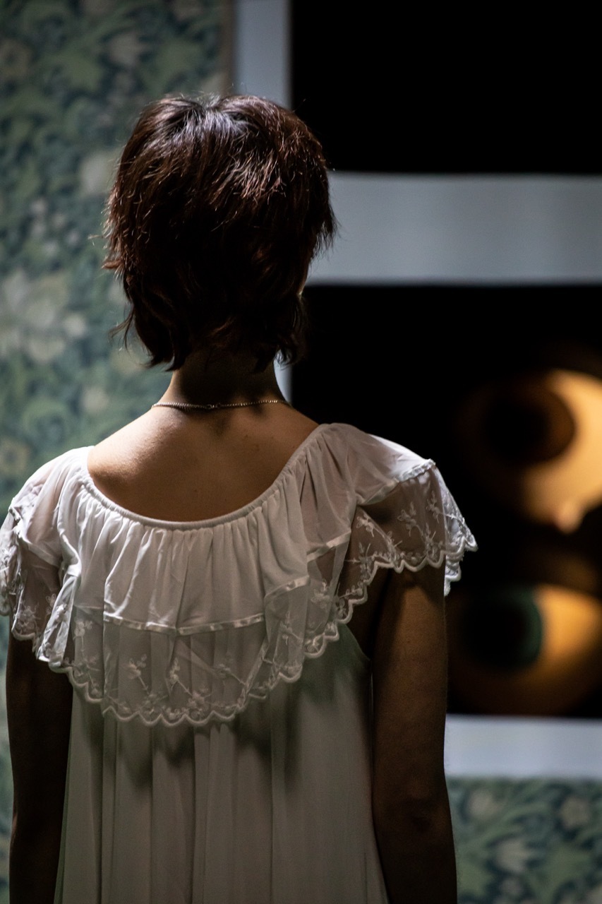 Woman with short brown hair viewed from behind wearing white ruffled blouse with lace trim, standing indoors with blurred background.