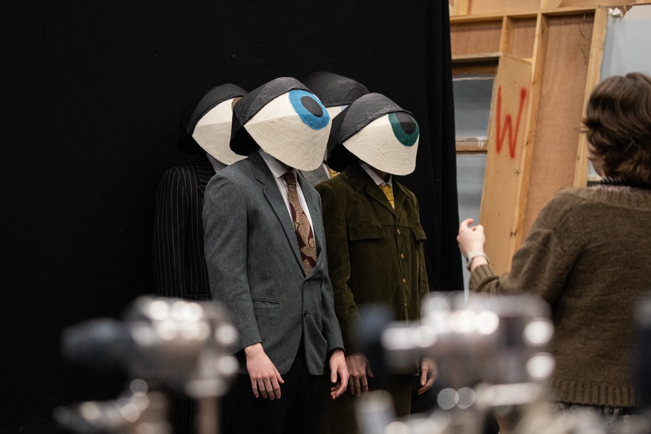 Three people in suits wearing large white masks with single blue and green circular eye designs, standing indoors.