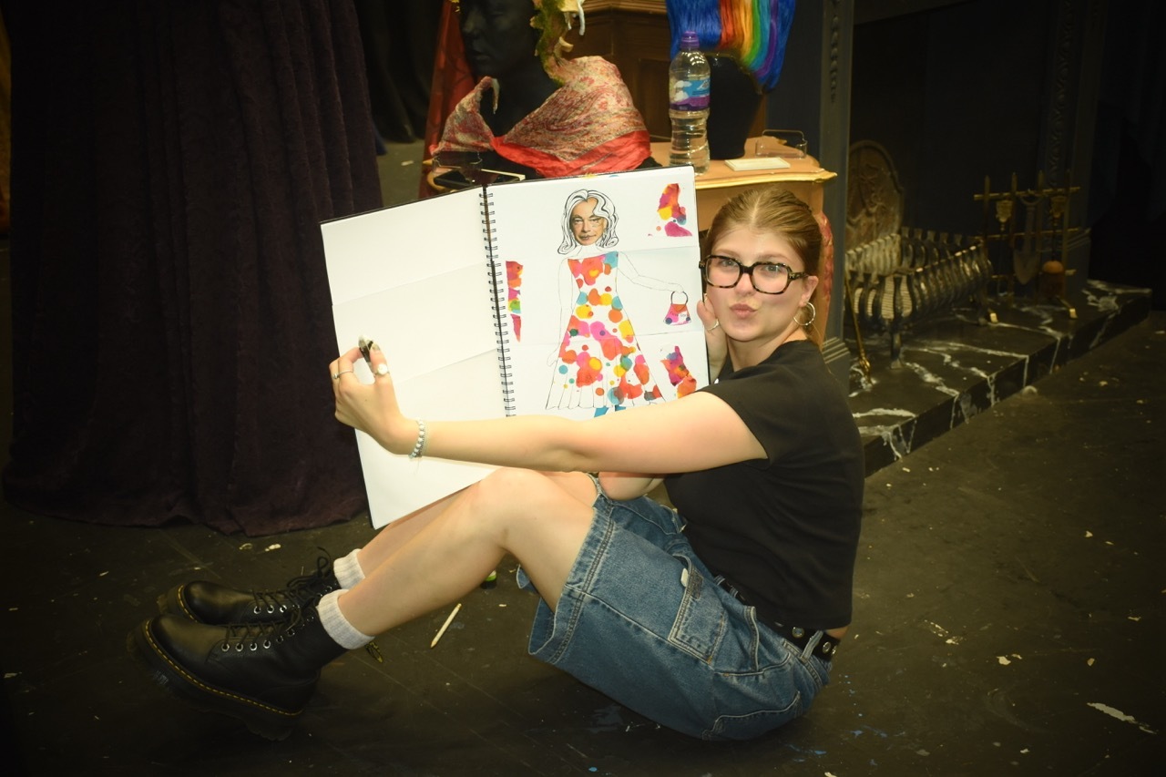 Woman with glasses sits on floor holding large sketchbook with colourful character drawings, surrounded by theatre props and costumes.
