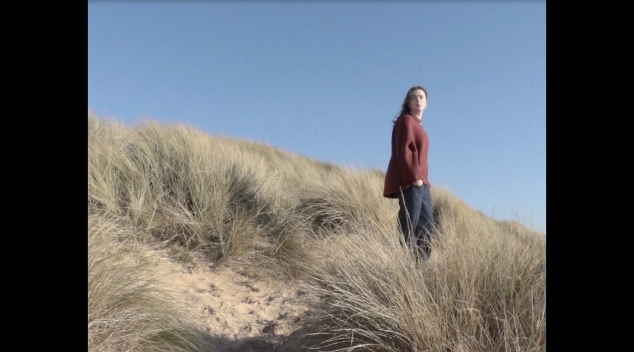 Woman in red top standing on sandy dune with tall golden grass against clear blue sky.
