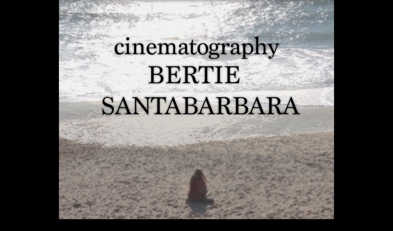 Film credit text "cinematography BERTIE SANTABARBARA" overlaid on desert landscape with single figure sitting on sandy ground.