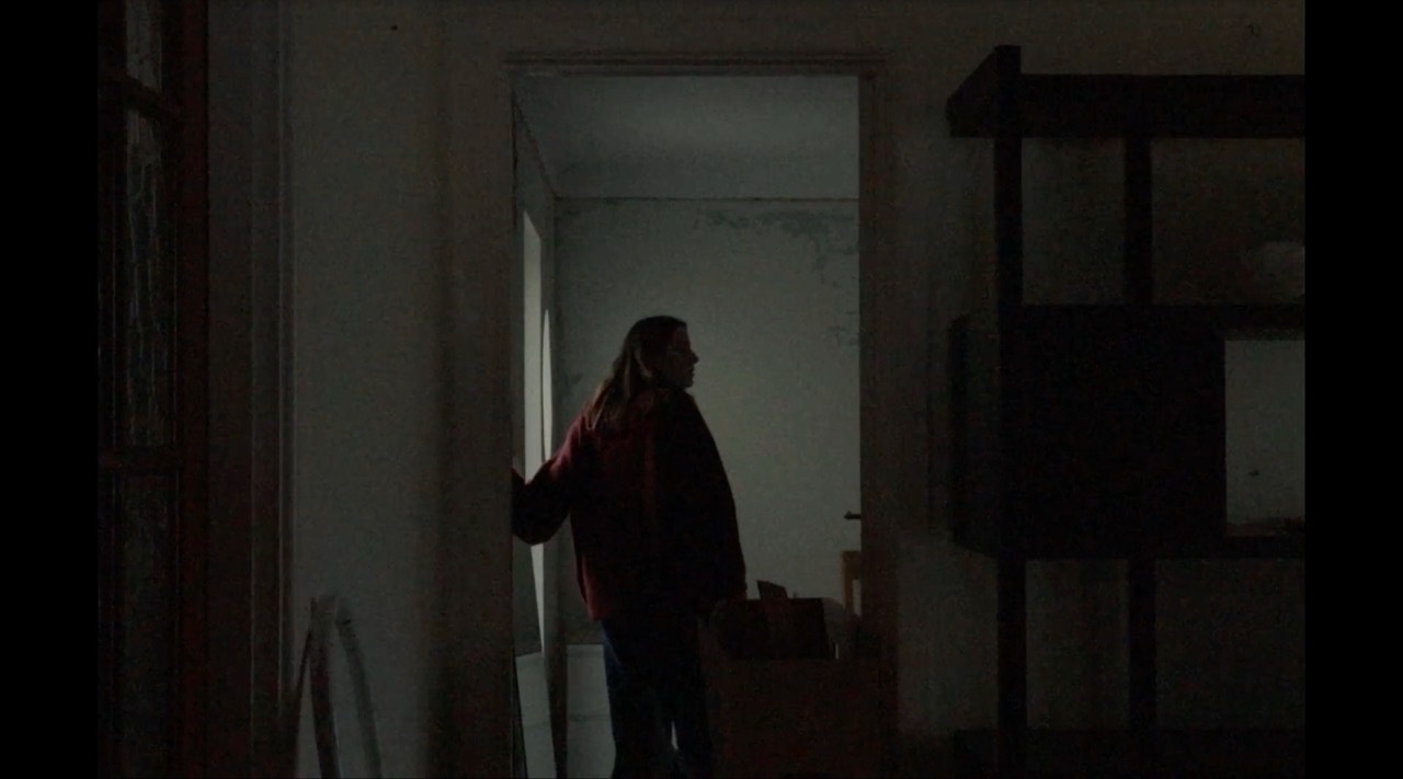 Person silhouetted in doorway between dark room and brightly lit corridor, creating strong contrast between shadows and light.