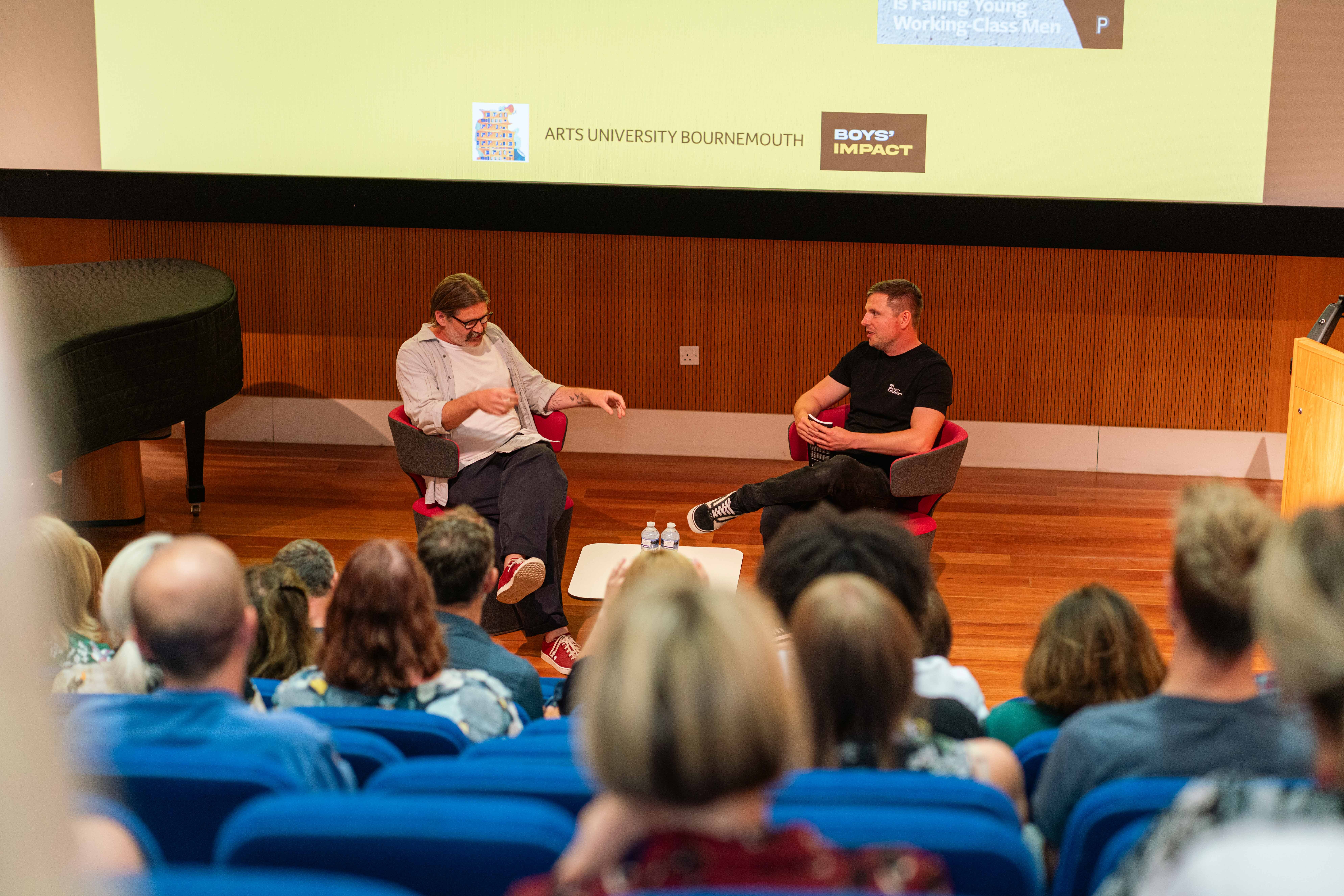 D Cox and Dr A. Blower seated on stage in chairs having discussion, audience in blue seats facing them, yellow wall with logos behind.
