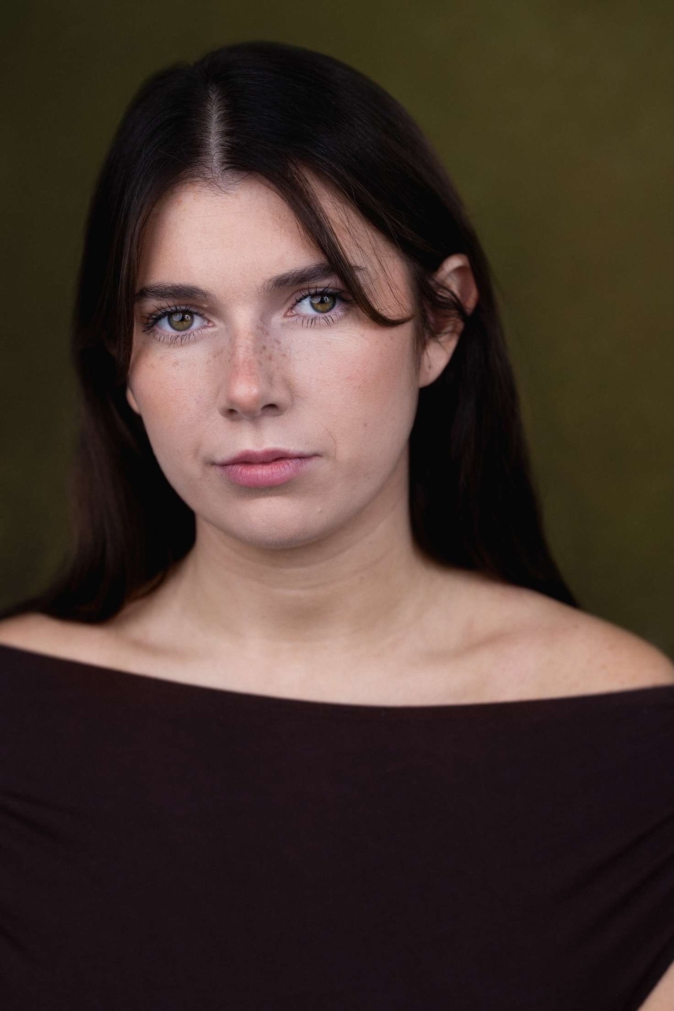 Woman with long dark hair wearing black off-shoulder top against olive green background, direct gaze at camera.