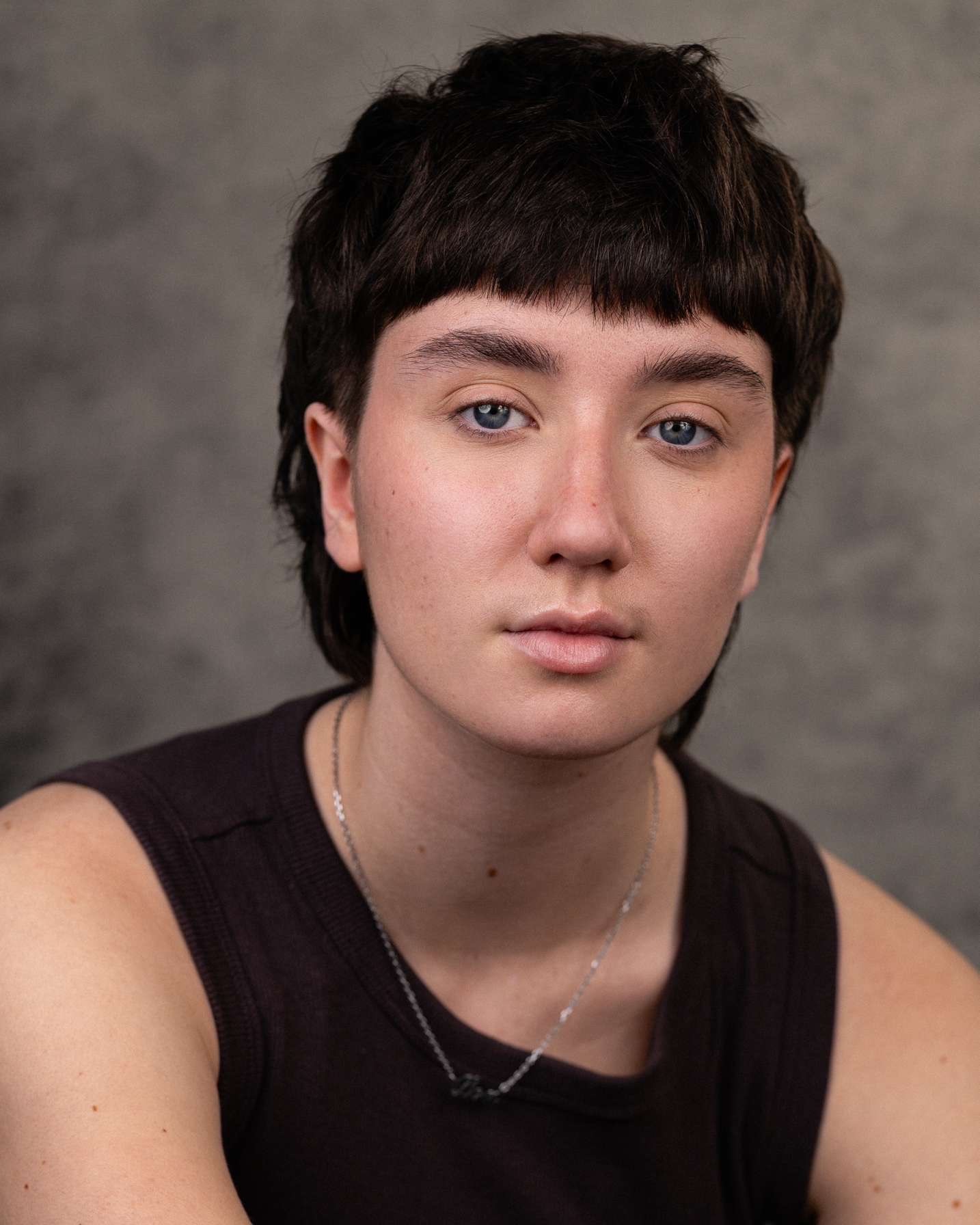 Person with short dark hair and fringe wearing black top, looking directly at camera against grey background.