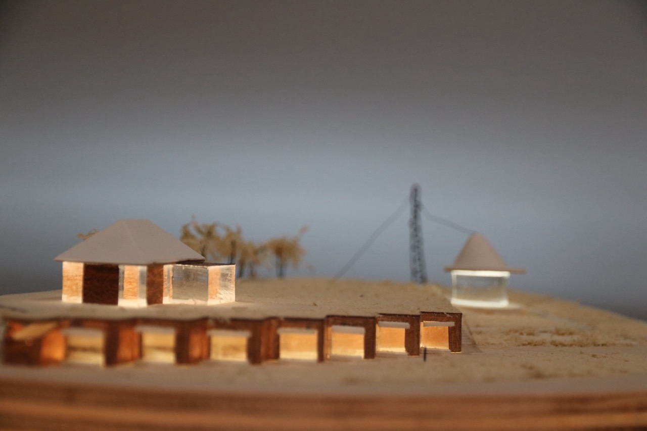 A small architectural model depicting two buildings with triangular roofs, one illuminated. In the background, a power line is visible, alongside wooden structures arranged in a line. The scene is set against a neutral grey background.