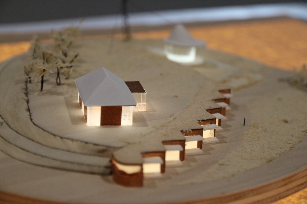 A scale model depicting two structures with sloped roofs on a sandy terrain. The model features a white building and a brown one, with illuminated sections, surrounded by trees and small wooden walkways leading towards the structures.