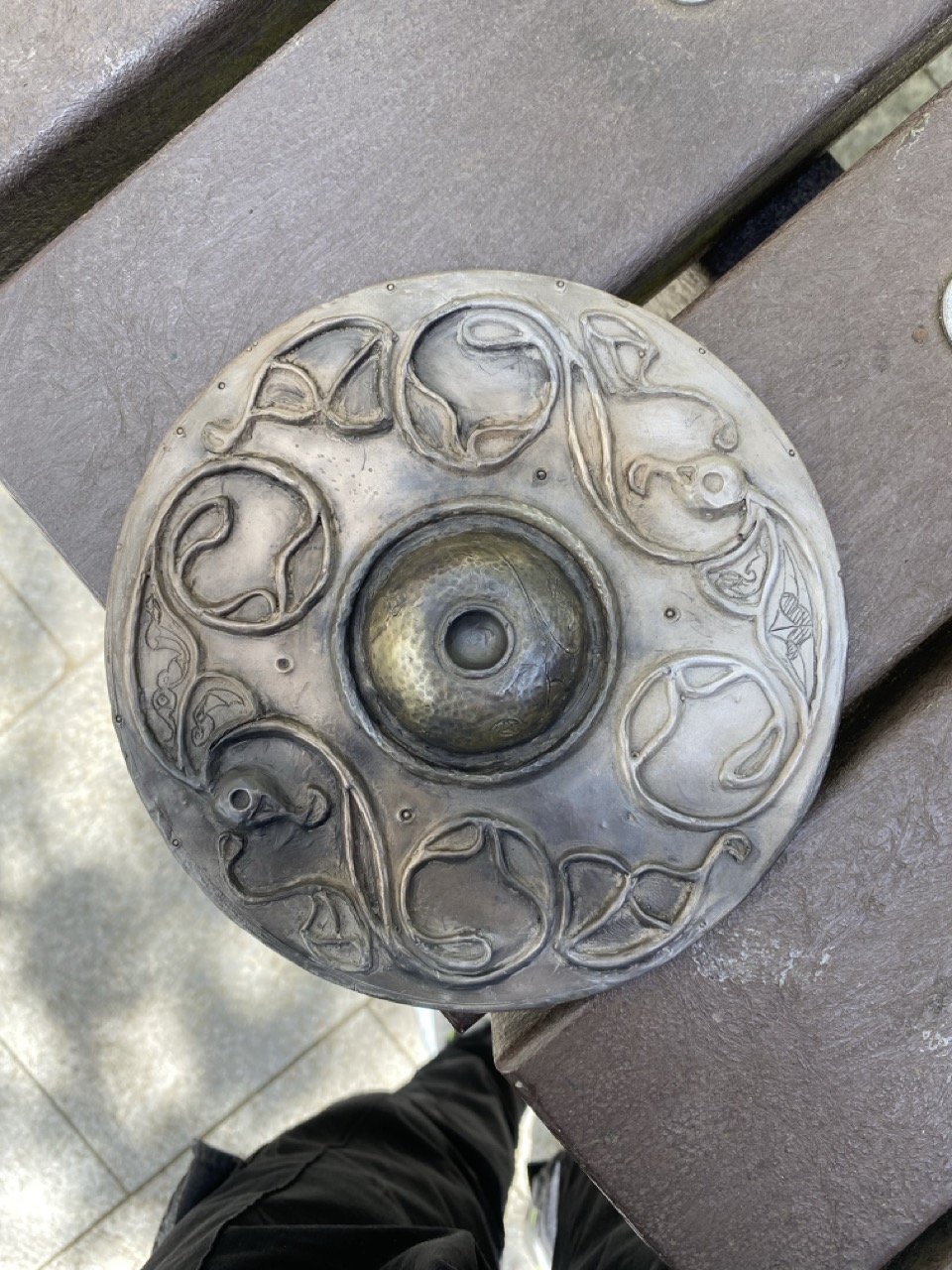 A circular, metal object with intricate swirling patterns and a central circular depression, resting on a wooden surface. The surface shows signs of wear, and part of a person's leg in dark clothing is visible at the bottom of the frame.