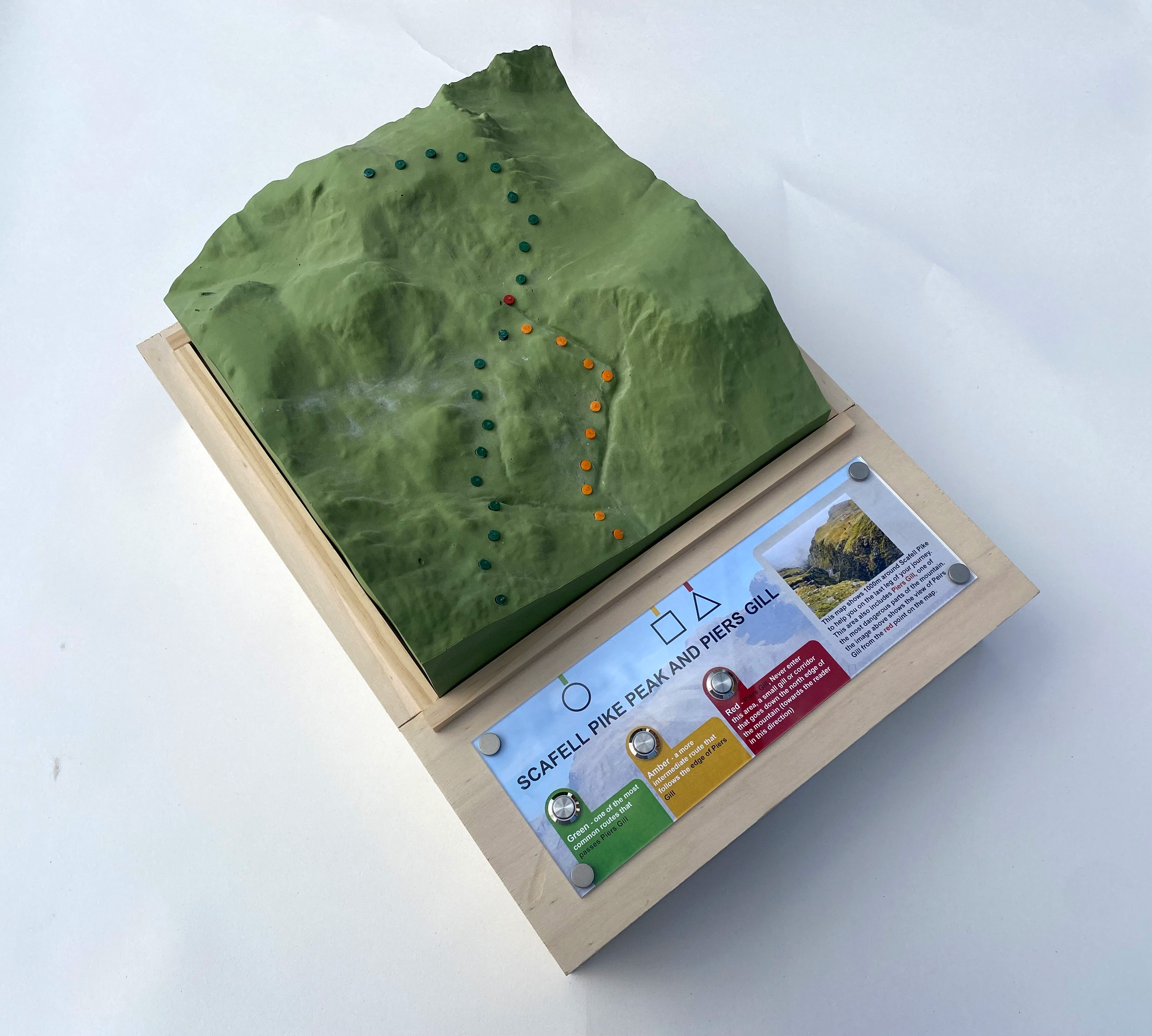 A topographical model depicting Scafell Pike and Piers Gill, with a textured green surface representing the terrain. Coloured dots indicate a walking route, and an information panel at the bottom details the area’s features and trail options.