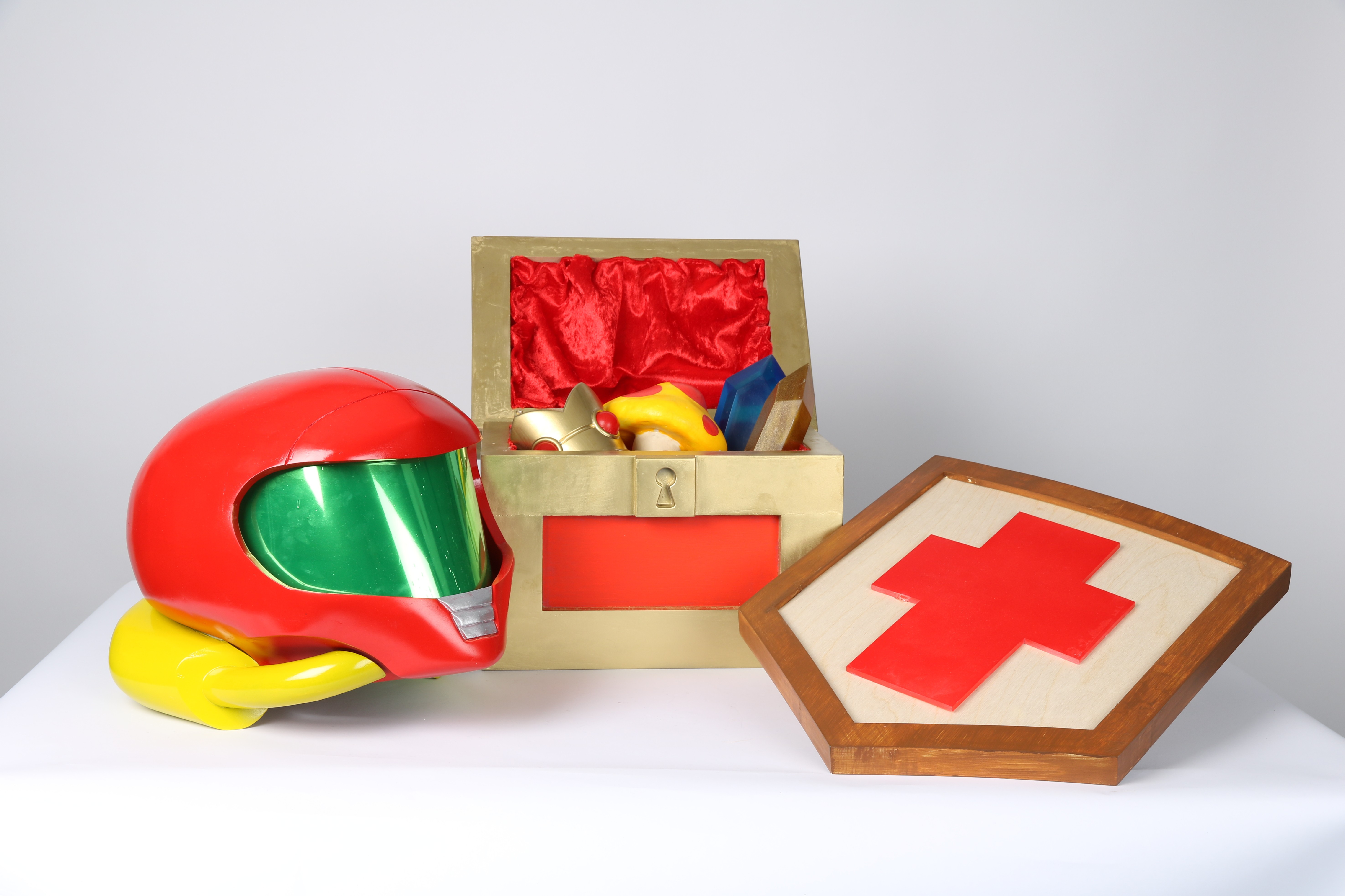 A bright red helmet with a green visor rests next to a golden treasure chest lined with red fabric, containing various toys. A wooden shield with a red cross design is also displayed prominently beside the helmet.