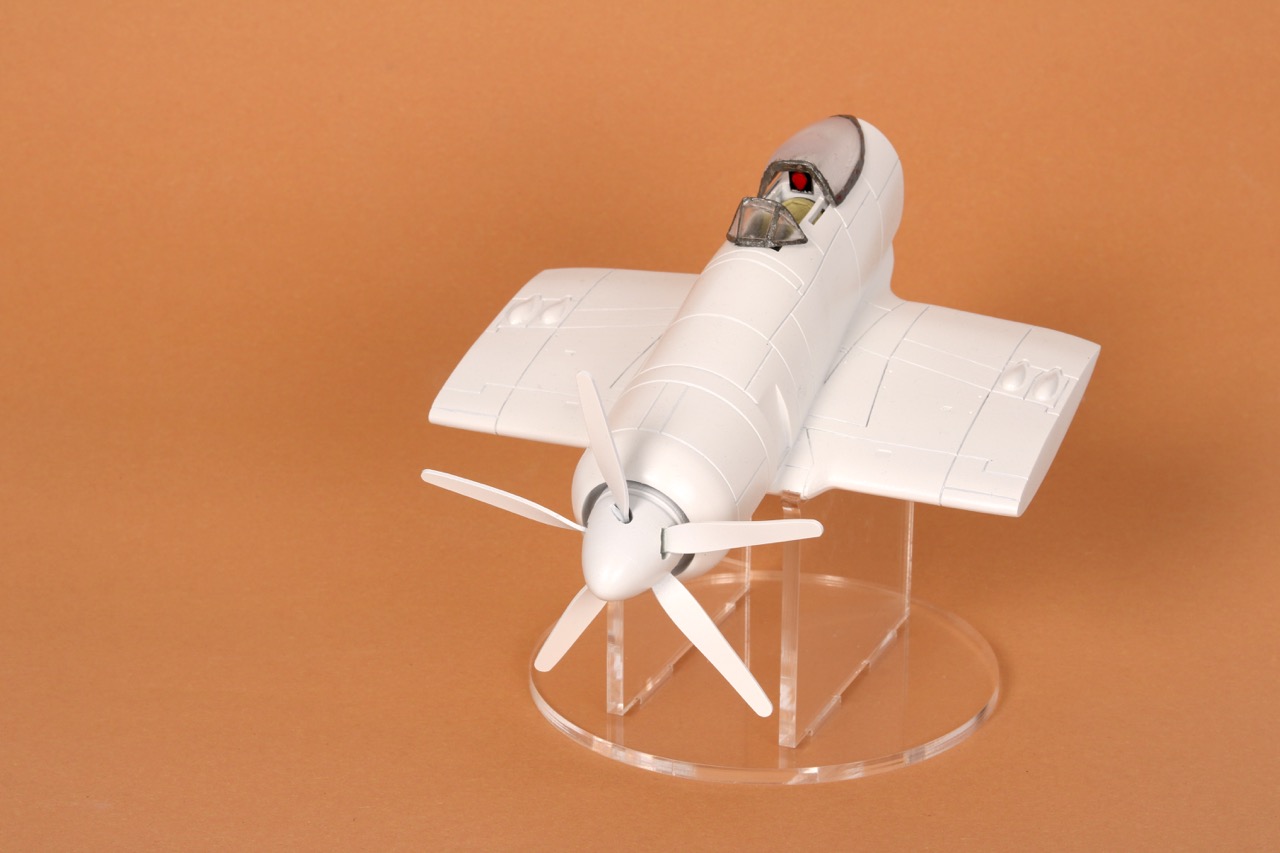 A white model aeroplane with a streamlined fuselage and four-blade propeller, displayed on a clear stand against a light brown background. The cockpit features a small figure of a pilot.