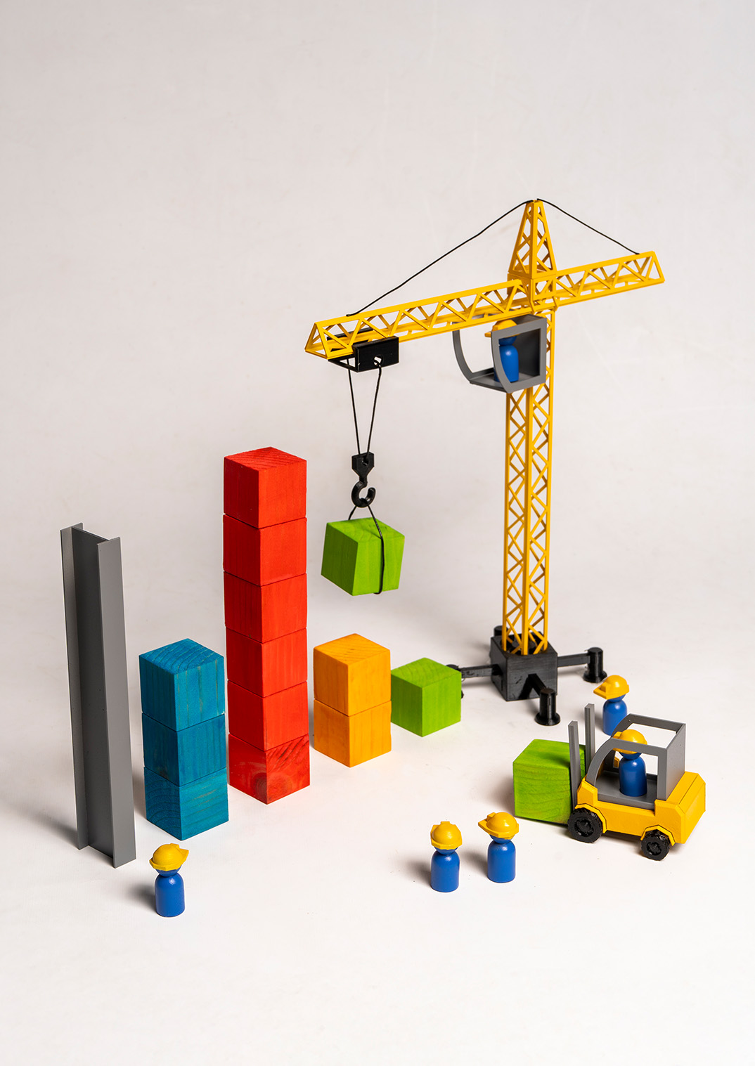 A construction toy scene featuring a yellow crane lifting a green box, alongside colourful stacked blocks forming bar graphs in red, blue, and orange. Two toy figures, one in a blue outfit and another in a yellow hard hat, stand near a yellow forklift.