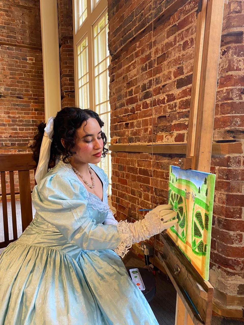Kasandra Georgi dressed in a Victorian-style light blue dress, painting a green landscape onto a small canvas, brick wall background.
