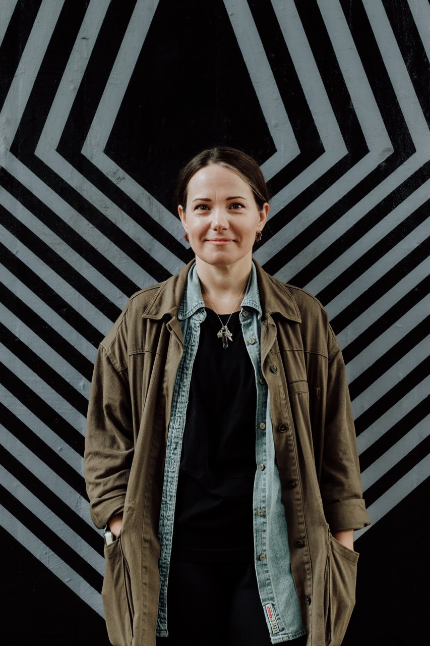 Louisa Flynn wearing a green coat stood in front of a black and white stripe background.