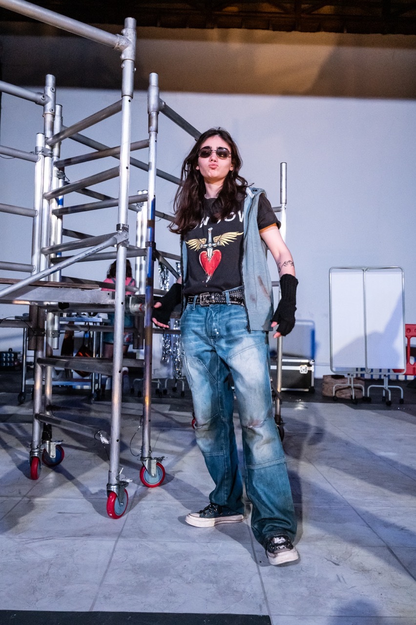 Woman in sunglasses, black graphic t-shirt, denim jacket and jeans stands beside metal scaffolding on wheels in industrial space.