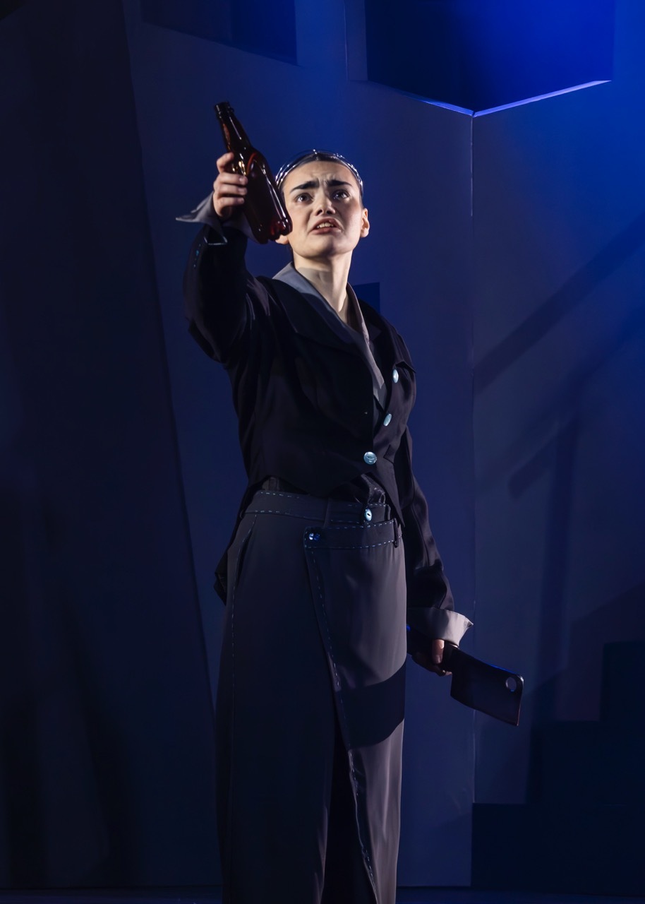 Woman in dark suit holding pistol aloft on stage with dramatic blue lighting and geometric backdrop.