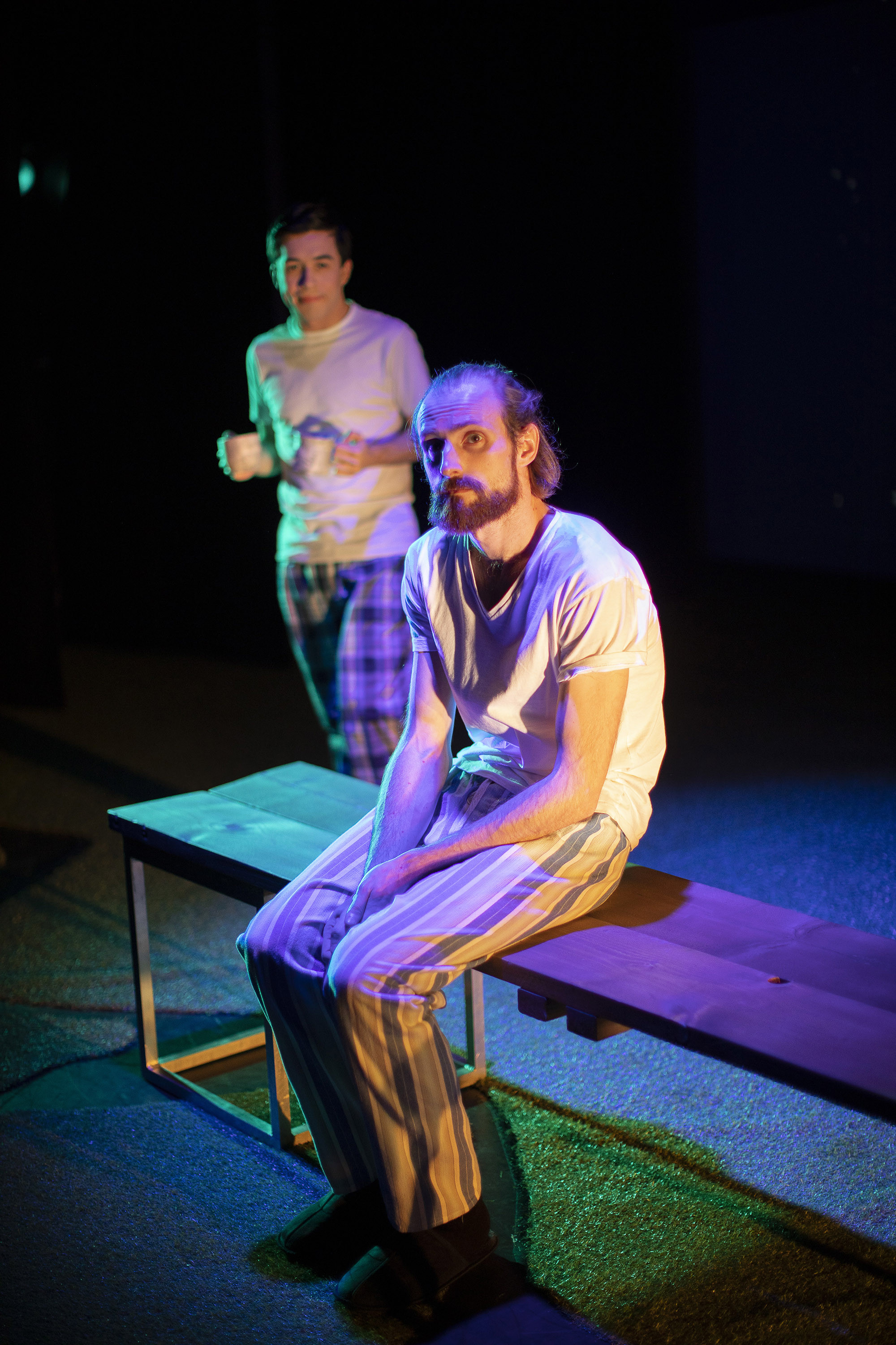 A person in pyjamas sitting on a bench on a stage. Another person, also in pyjamas, approaches from behind with coffee mugs in their hands.
