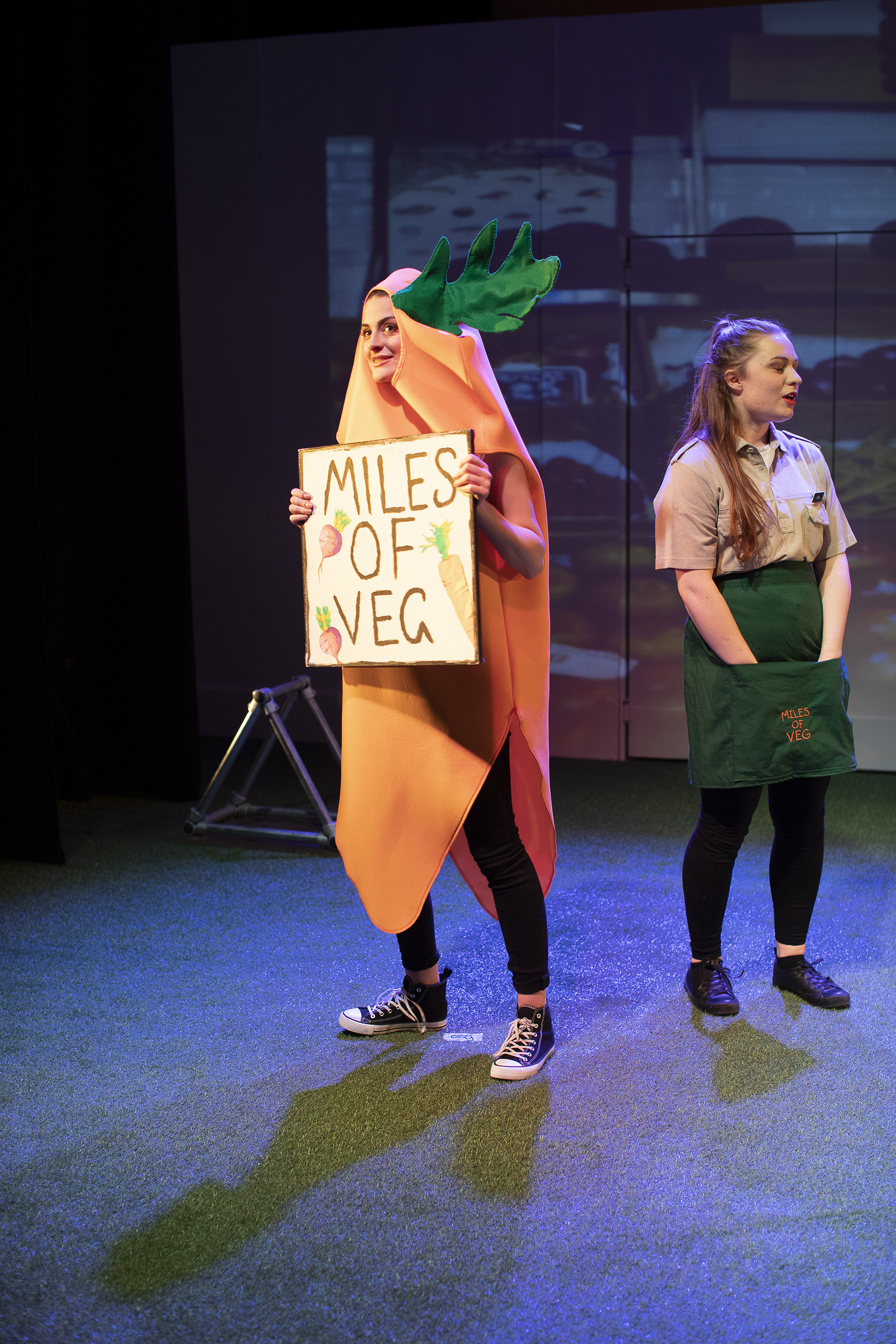 Two people standing on a stage. One is dressed as a carrot holding a sign reading "Miles of veg". The other person is wearing an apron and has their hands in their pockets.