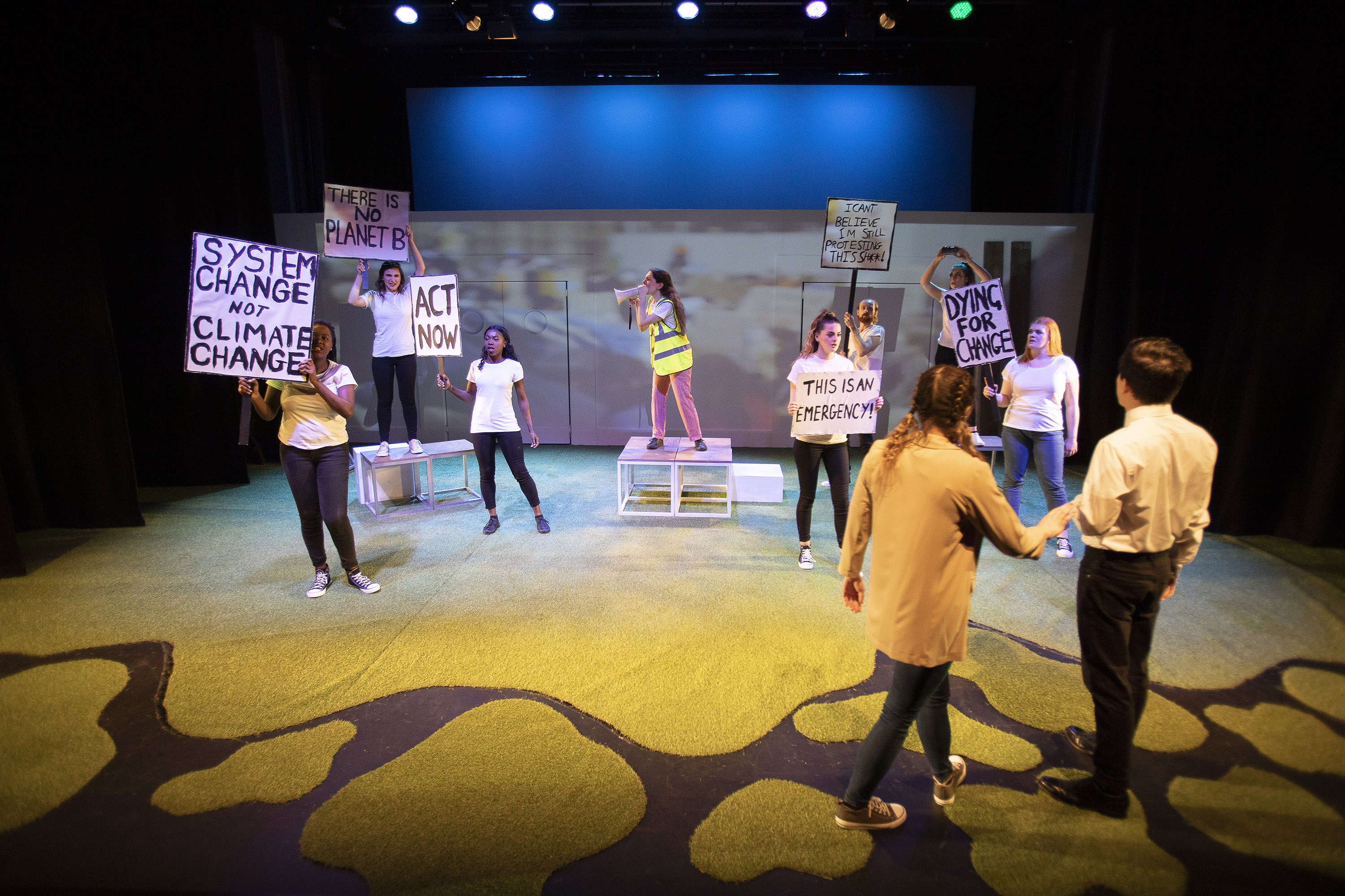 A protest scene on stage with actors standing in a circle and holding placards. A person in a hi-vis jacket is standing on a block in the centre and speaking into a loudspeaker.