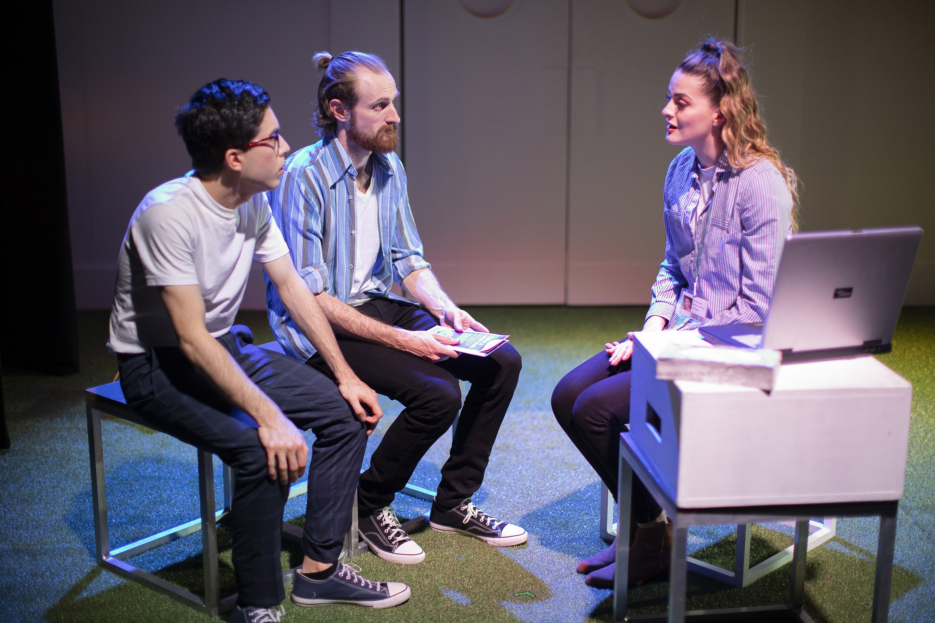 Three people sat on a stage talking, two are seated on a bench on the left, the other person sits opposite them with a laptop open in front of them.