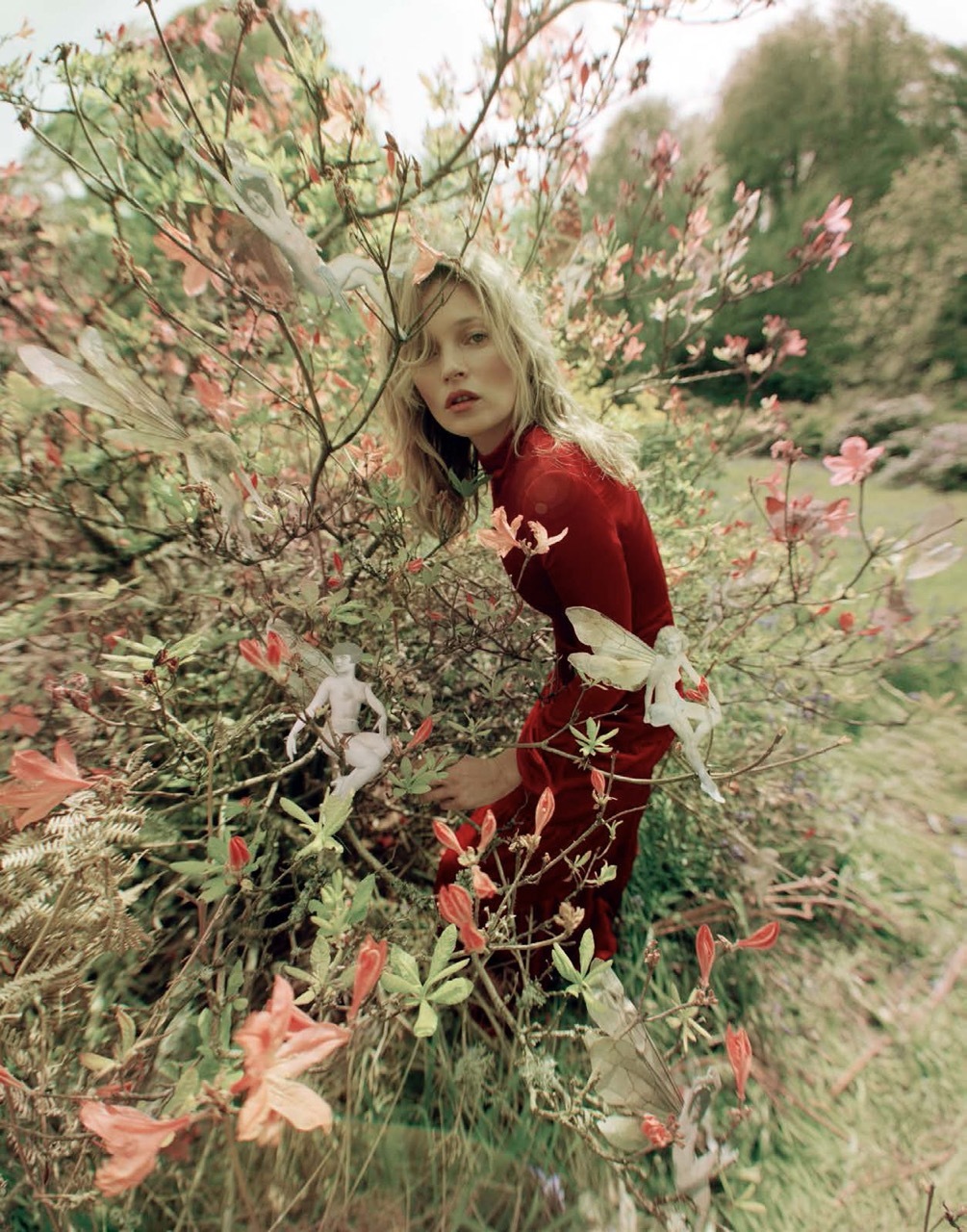 A woman wearing a red dress amongst a bush and flowers.