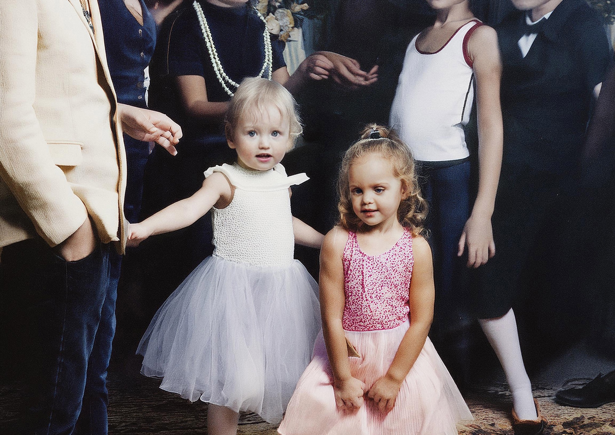 Two children wearing party dresses. Adults and other young people are visible in the background.