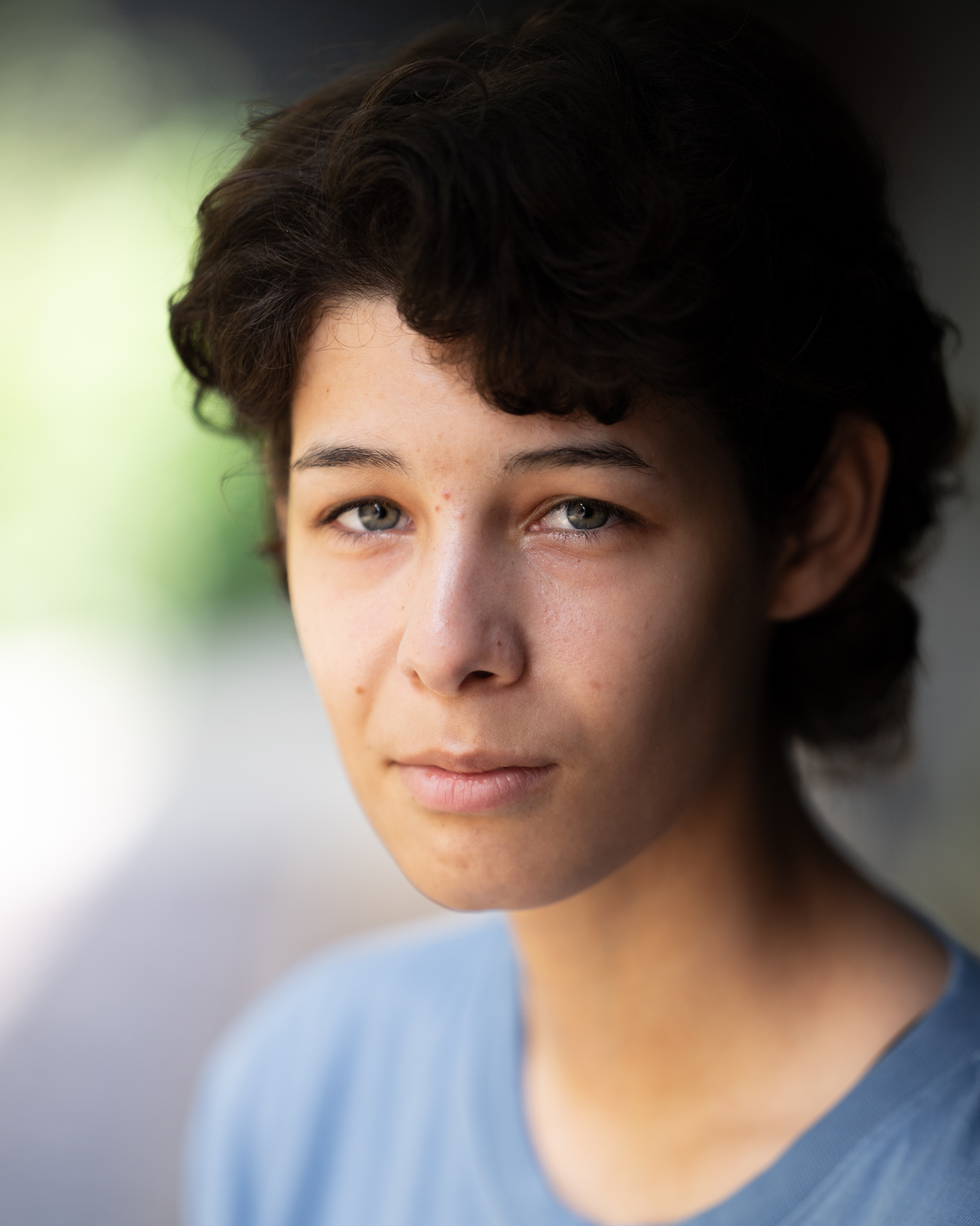 Young person with short curly brown hair wearing light blue clothing, soft natural lighting, blurred background.