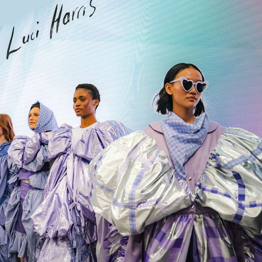 Models in oversized metallic silver and purple garments against gradient blue-purple background. "Luci Harris" text in top left corner.