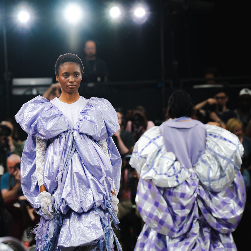 Models on runway wearing oversized lilac ruffled dress with voluminous sleeves, bright stage lights above, audience visible in background.