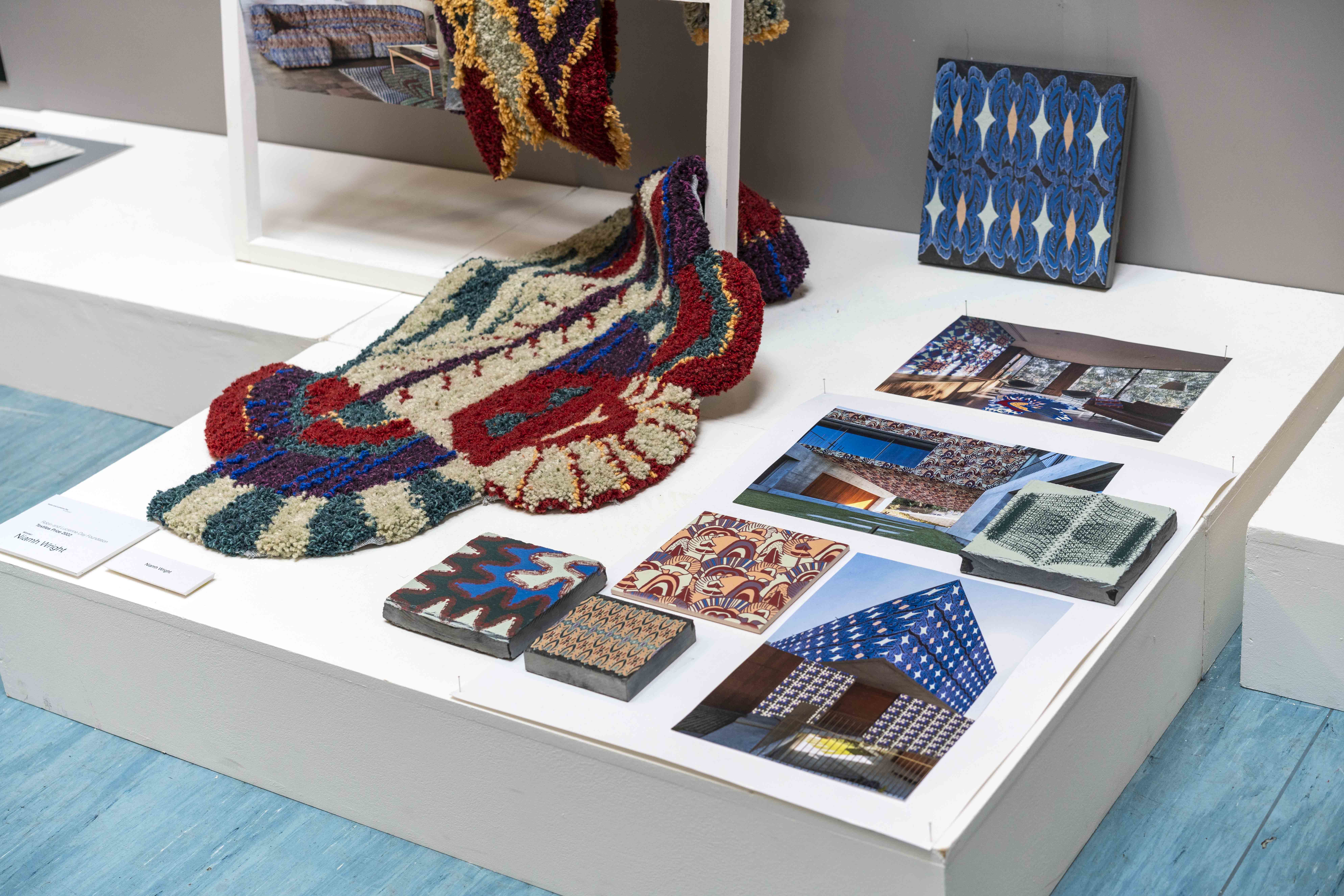 A display of canvases and a large colourful rug lie on a plain surface in a gallery space