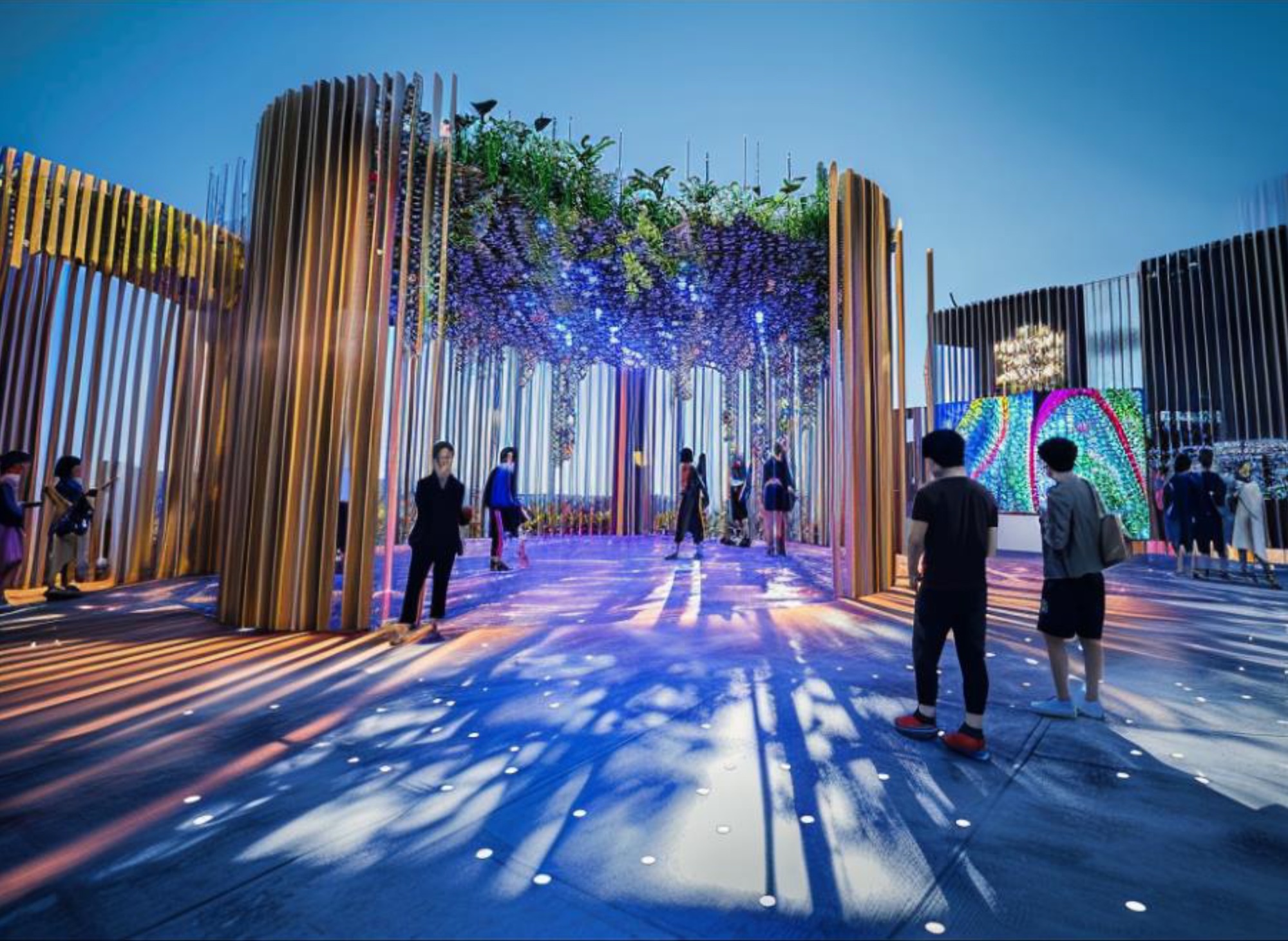 Modern installation with tall wooden slats, hanging greenery, blue lighting projected on floor, and visitors exploring the space.