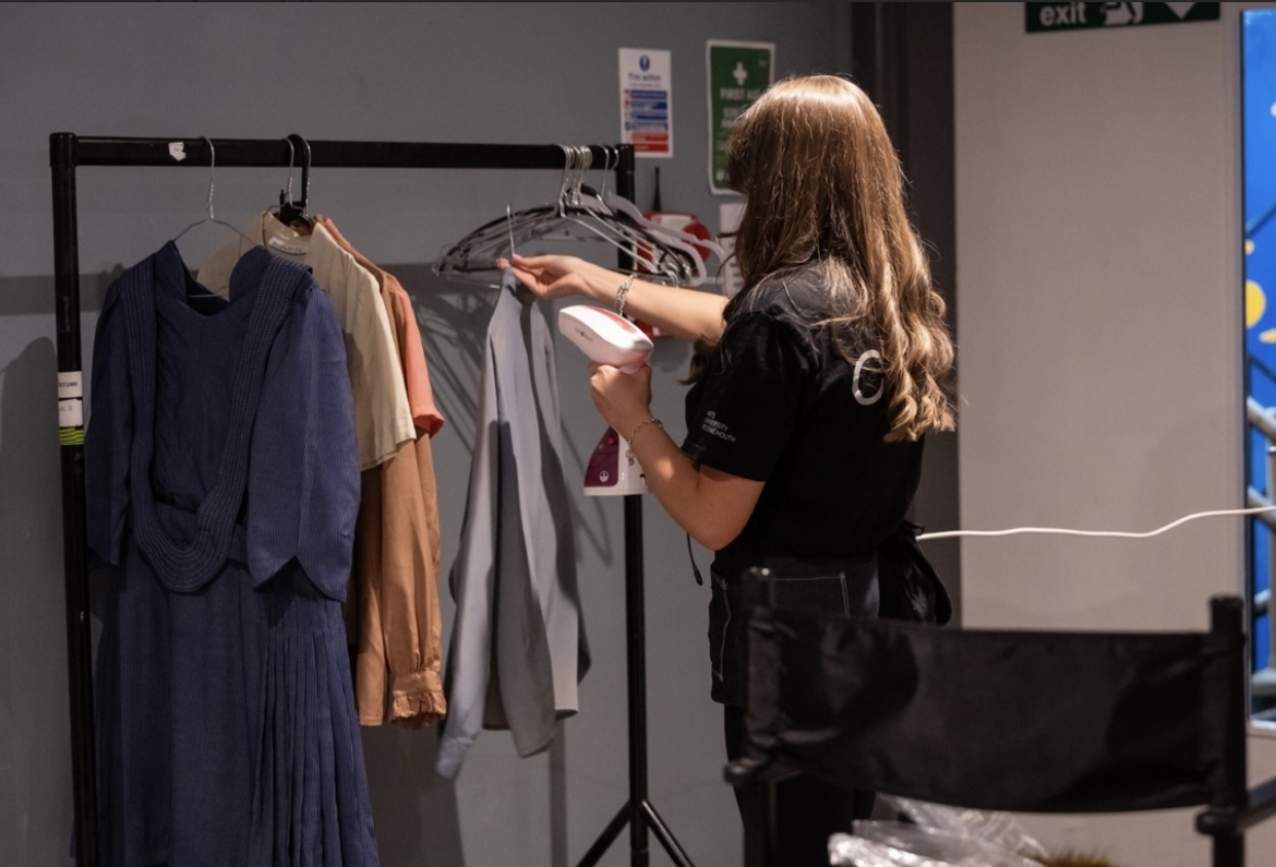 Woman with long brown hair browsing clothes on black metal rail in shop, wearing dark jacket with handbag.