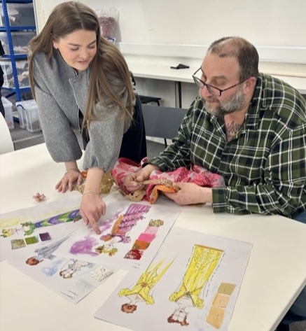 Woman in grey jumper and bearded man in green checked shirt examining colourful drawings spread on white table in classroom setting.
