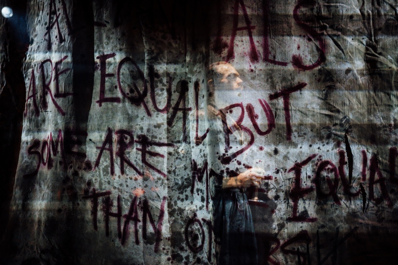 A person stands behind a graffiti-covered wall with the words “ARE EQUAL BUT...” painted in red and black.