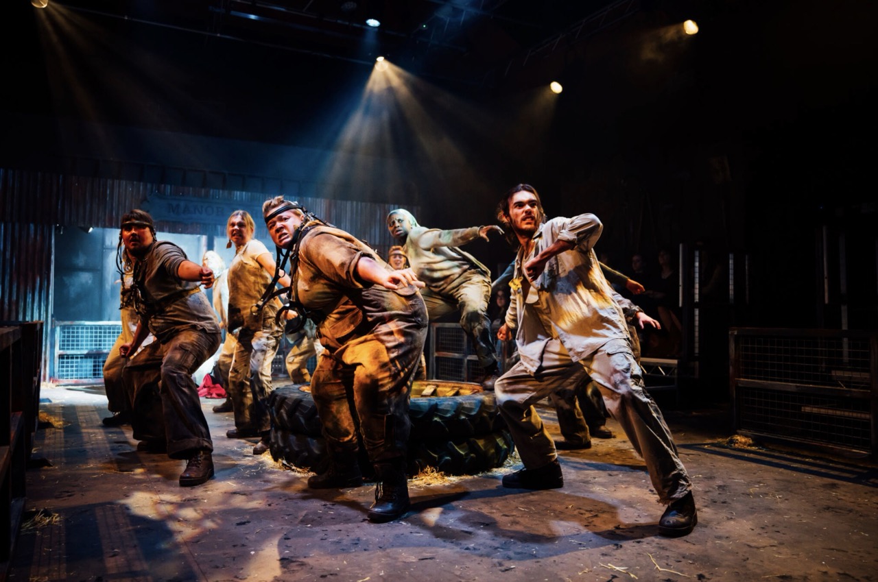 Four performers in distressed workwear pose around a person on the floor under stage spotlights, with a dark industrial set and metal fencing in the background.